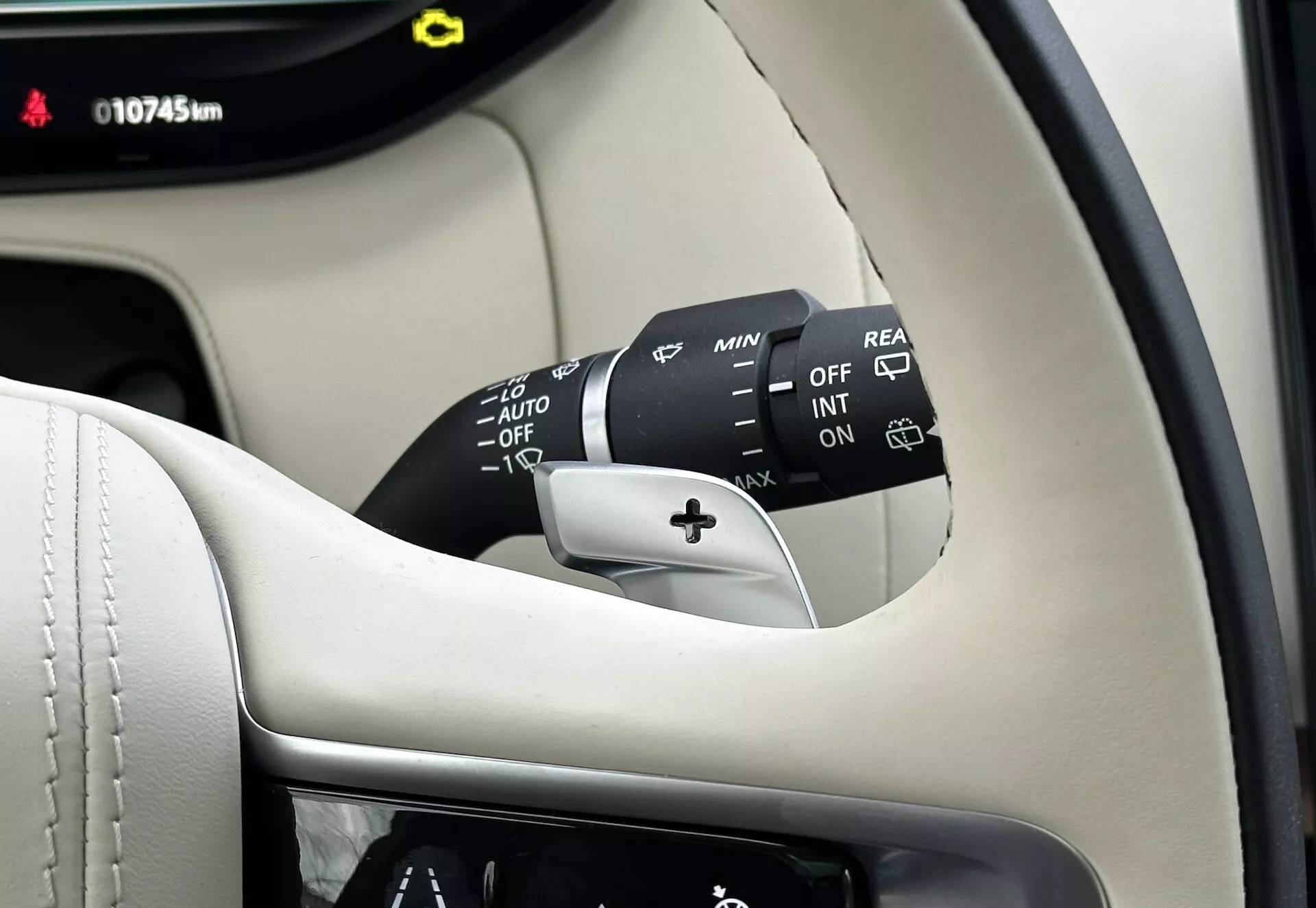 Vista ravvicinata dell'interno della Land-Rover Range-Rover 2021 che mostra la colonna del volante, la leva di controllo del tergicristallo con molteplici impostazioni inclusi OFF, INT, ON, regolatori di velocità e AUTO per tergicristalli automatici con sensore di pioggia, più un'opzione per il tergicristallo posteriore. Parte visibile del cruscotto mostra 10745 km, ambientata contro un interno premium in nero e crema.