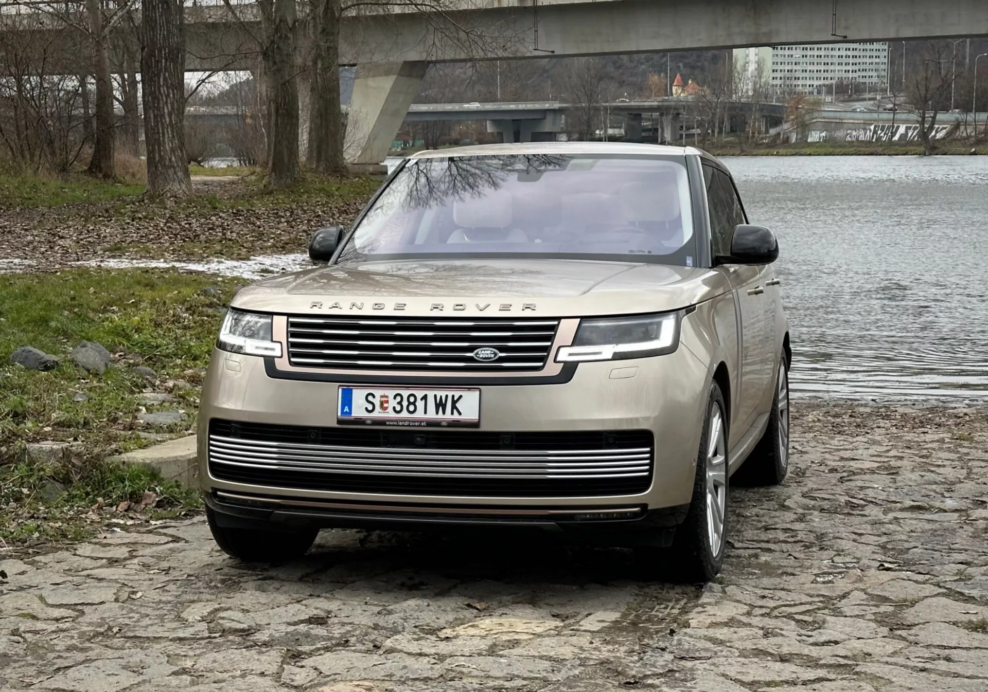 SUV di lusso Range Rover 2021 in argento, mette in mostra la griglia frontale prominente con il branding, luci diurne LED eleganti, paraurti anteriore con sensori di parcheggio e design del cofano clamshell su una superficie acciottolata vicino a un fiume