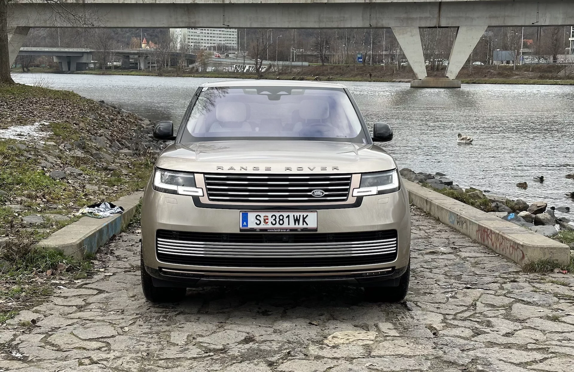 Land Rover Range Rover 2021 fotografato da un angolo frontale e leggermente basso, che evidenzia la griglia, il paraurti anteriore e il cofano, con l'iconico emblema RANGE ROVER, fari LED moderni e tecnologia avanzata di assistenza alla guida, su sfondo di un corpo d'acqua con un ponte e un cielo nuvoloso