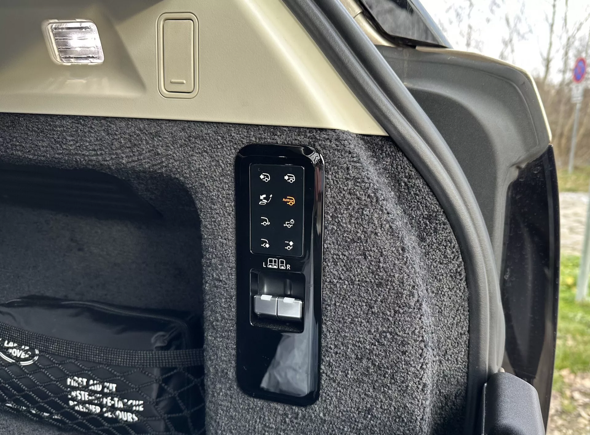 Vista ravvicinata del pannello di controllo elettronico del bagagliaio interno di una Land Rover Range Rover 2021, mostrando pulsanti per operazioni come apertura e chiusura elettronica, controllo del copri-carico, potenzialmente un luce del bagagliaio o presa di corrente, regolazioni dei sedili posteriori dal bagagliaio e rilascio di emergenza, impostato contro il lato del bagagliaio tappezzato e il soffitto chiaro con apparecchio di illuminazione del carico.