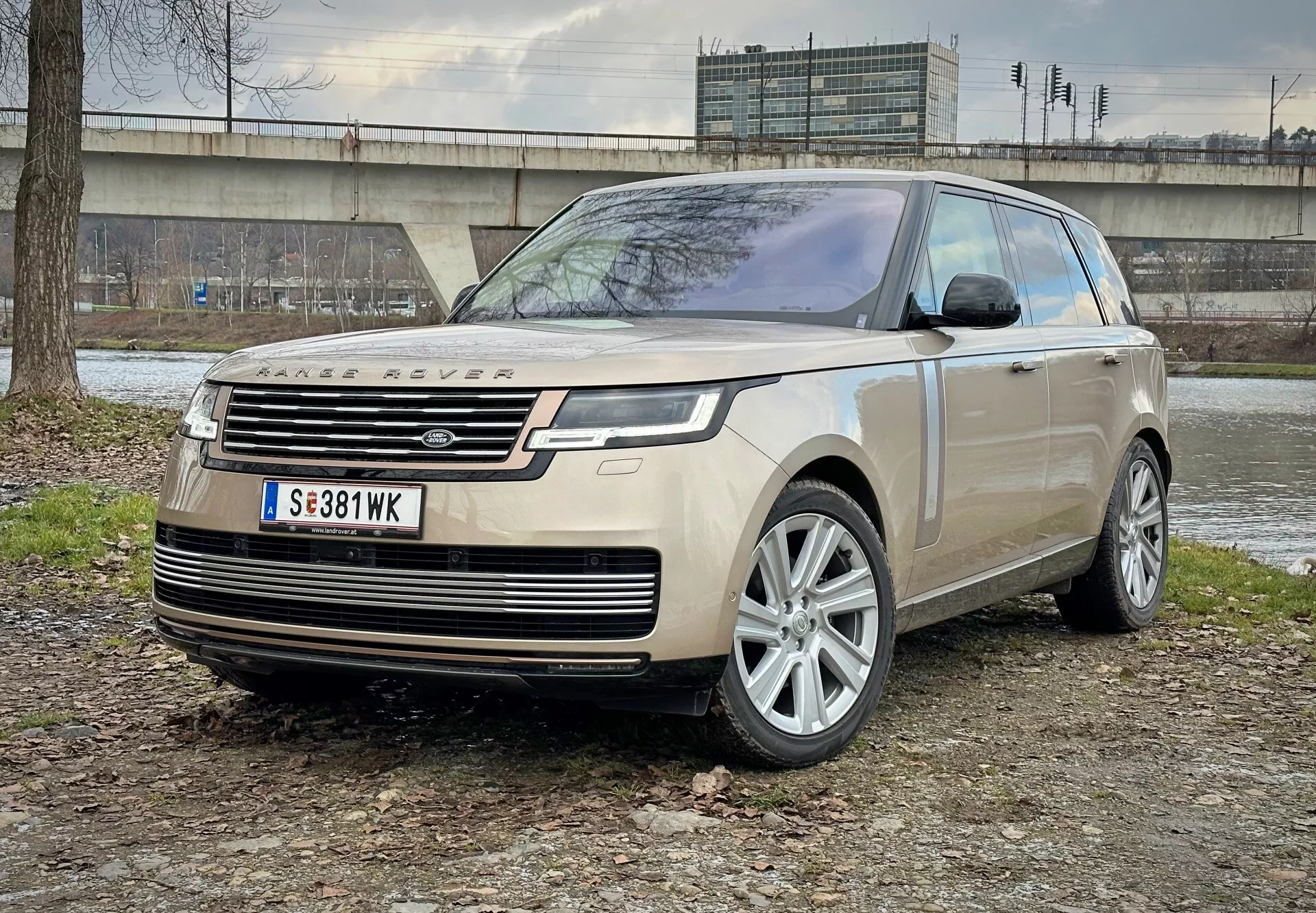 SUV di lusso Land Rover Range Rover 2021 parcheggiata su terreno misto di terra e erba con una struttura in cemento e vegetazione sullo sfondo, mostrando la vista da angolo anteriore evidenziando fari LED, ruote in lega e caratteristiche aerodinamiche moderne