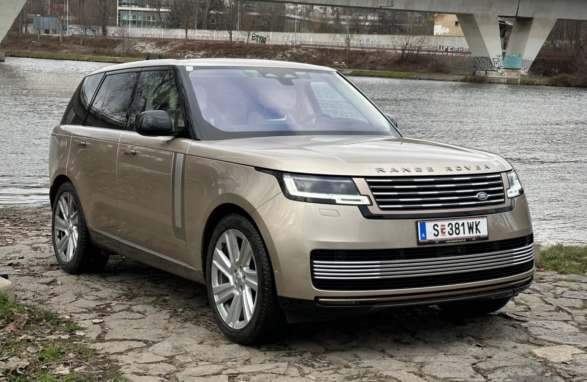 SUV di lusso Land Rover Range Rover 2021 parcheggiata in riva al fiume con un ponte sullo sfondo, che mette in mostra il suo design moderno e le caratteristiche tecnologiche tra cui fari LED, griglia frontale con emblema, cerchi in lega a più razze e tetto panoramico.