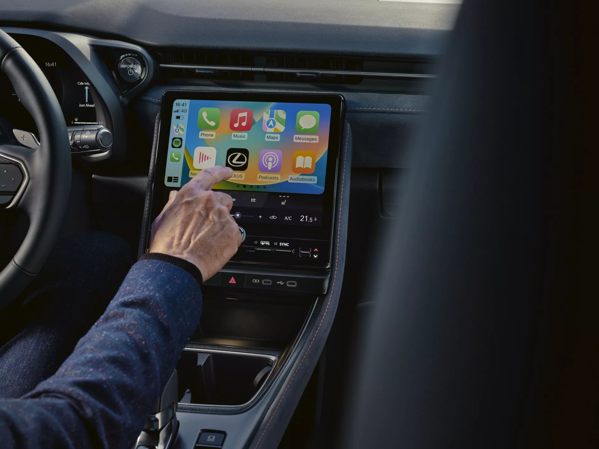 Vista ravvicinata dell'interno di una Lexus LBX 2024 con focus sul sistema di infotainment dalla prospettiva del passeggero, mostrando app di connettività e impostazioni del controllo clima su uno schermo touch verticale