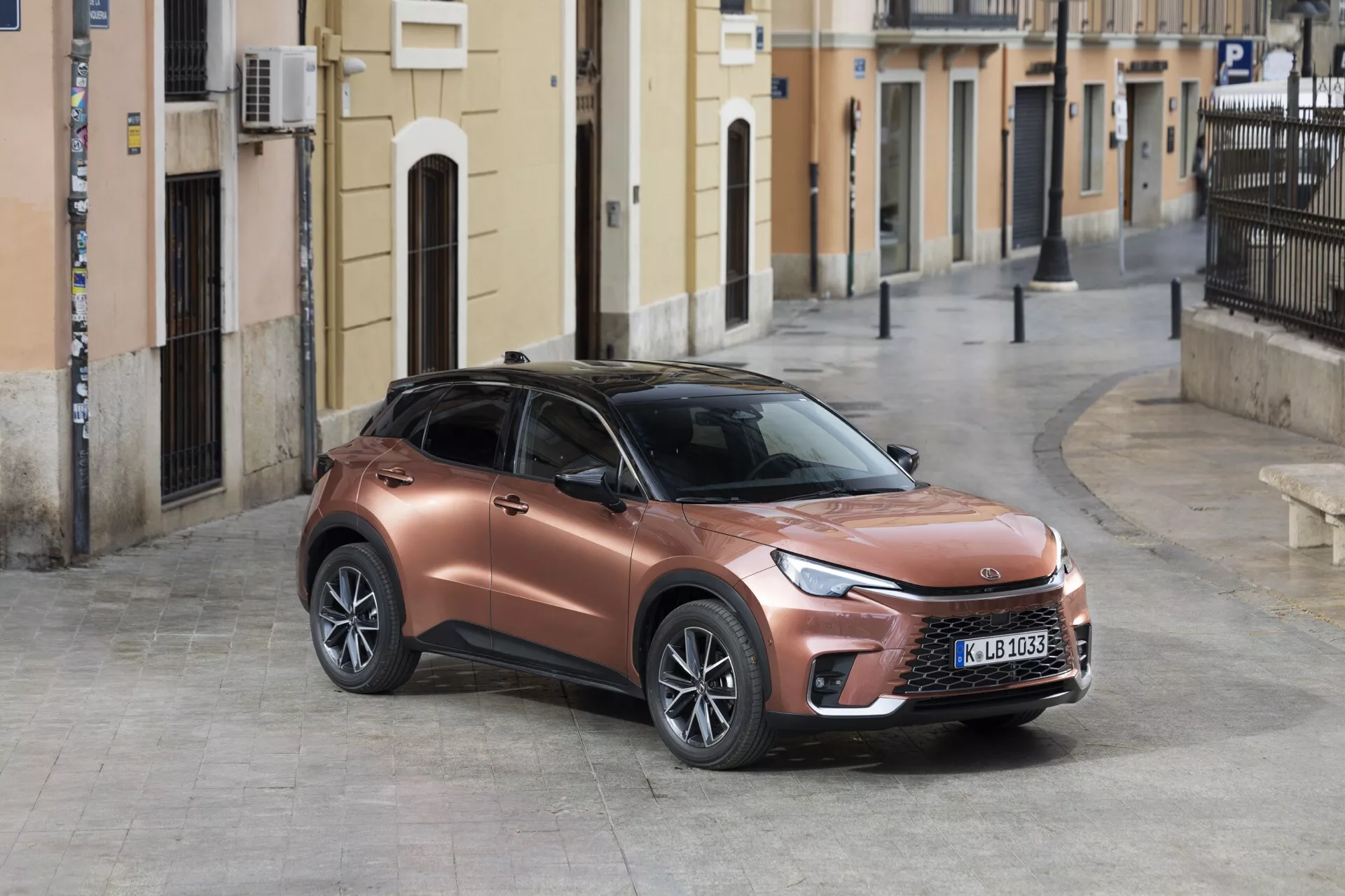 SUV crossover moderno Lexus LBX 1ª generazione 2024 parcheggiato su una strada acciottolata con vista a tre quarti dal lato anteriore sinistro che mette in mostra la sua finitura metallica bronzo, fari LED snelli e design sportivo rialzato