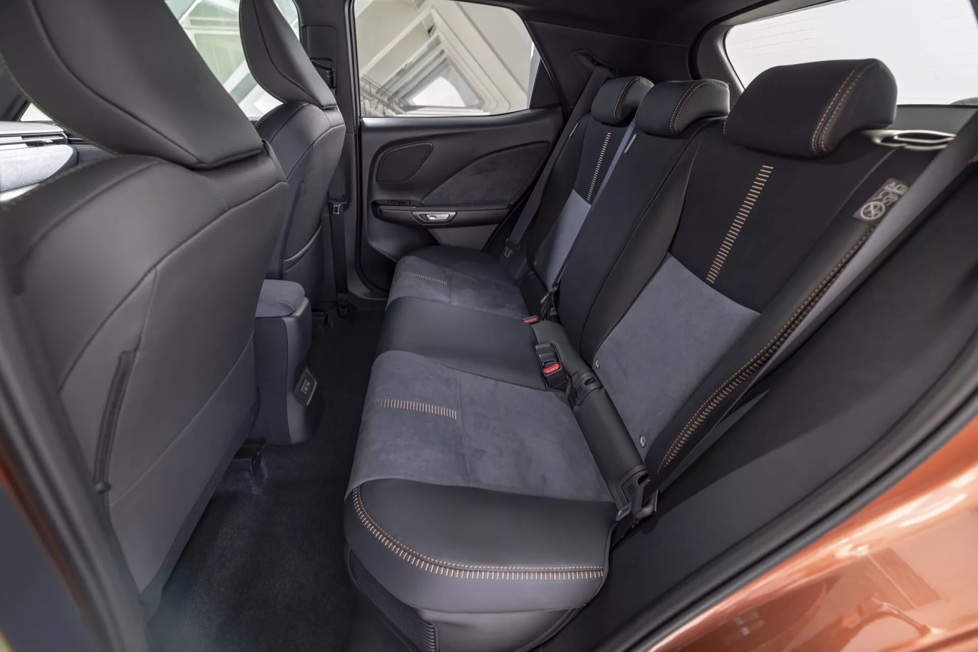Vista interna del Lexus LBX 1ª generazione 2024 dal comparto passeggeri posteriore che mostra l'area del sedile posteriore, dettagli della tappezzeria con cuciture a contrasto bianco, schienali trapuntati, bracciolo ribaltabile, griglia dell'altoparlante sulla porta e tasca di stoccaggio sul retro del sedile anteriore