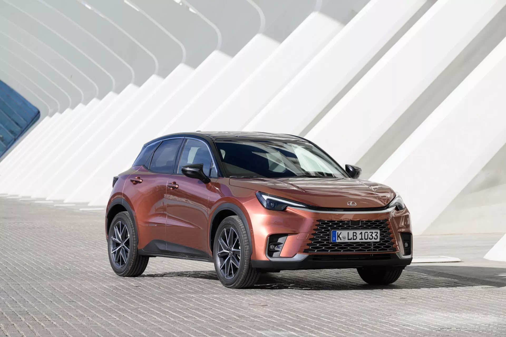 Lexus LBX di prima generazione 2024 esposta alla luce del giorno, caratterizzata da una griglia anteriore distintiva e fari LED eleganti, con uno sfondo di architettura moderna costituito da forme geometriche angolari. L'angolazione evidenzia la carrozzeria aerodinamica dell'auto, la vernice metallica e le ruote in lega, enfatizzando il suo design da SUV crossover e l'avanzata ingegneria automobilistica.