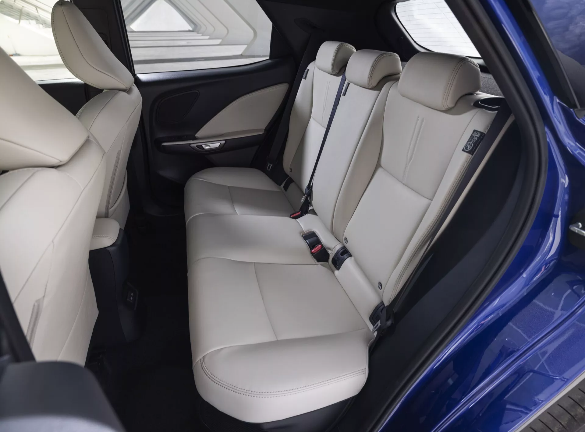 Interno del Lexus LBX 1ª Generazione 2024 che presenta sedili posteriori in pelle di beige chiaro premium con poggiatesta, cinture di sicurezza, pannello porta con comandi, sezione posteriore della console centrale inclusi bocchette d'aria e porte di ricarica, in condizioni di illuminazione ottimale che sottolineano confort e sicurezza