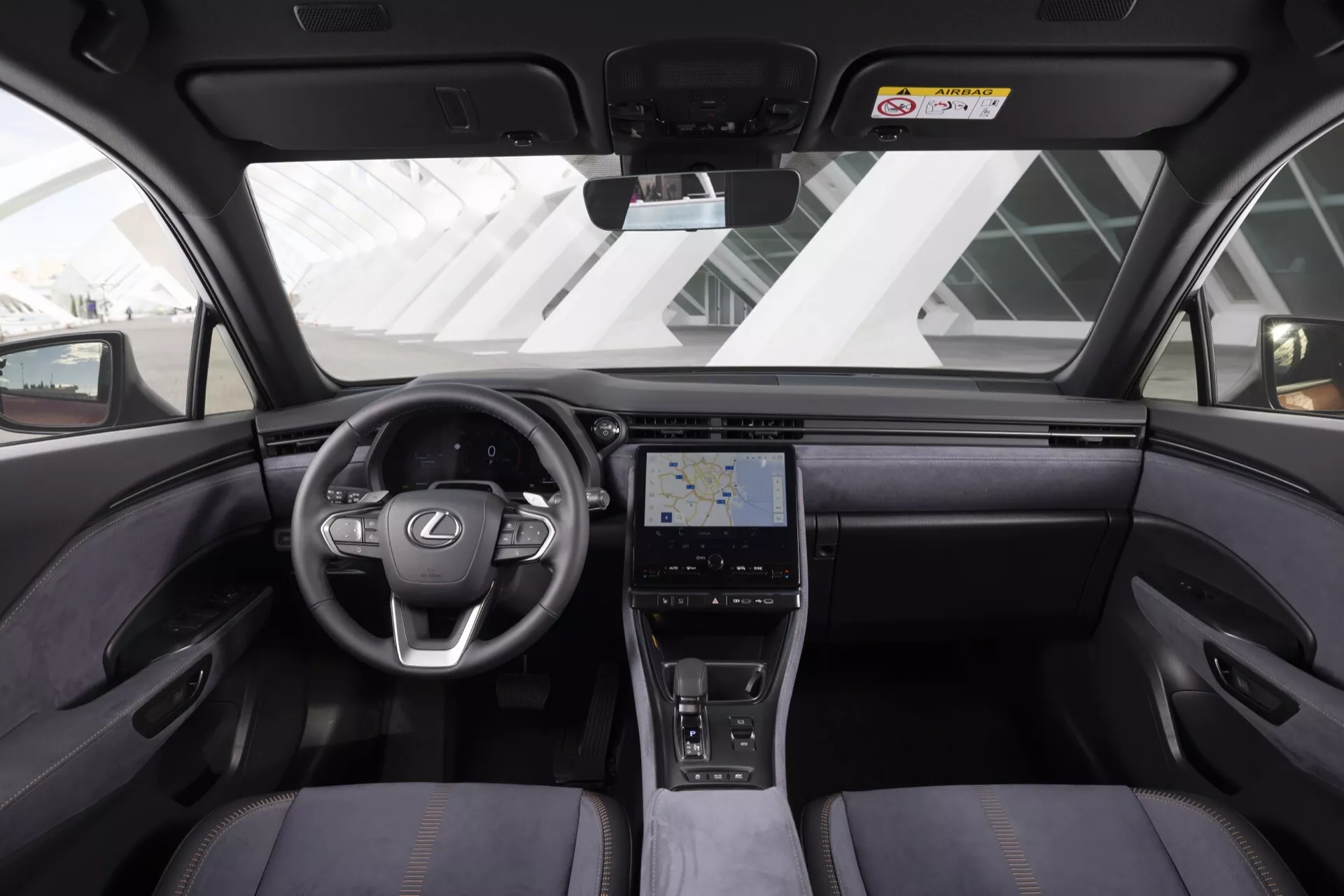 Vista interna della Lexus LBX 2024 che mostra cruscotto, volante con logo Lexus e controlli integrati, console centrale touch, sedili di lusso, e funzionalità tecnologiche avanzate dalla prospettiva del sedile posteriore
