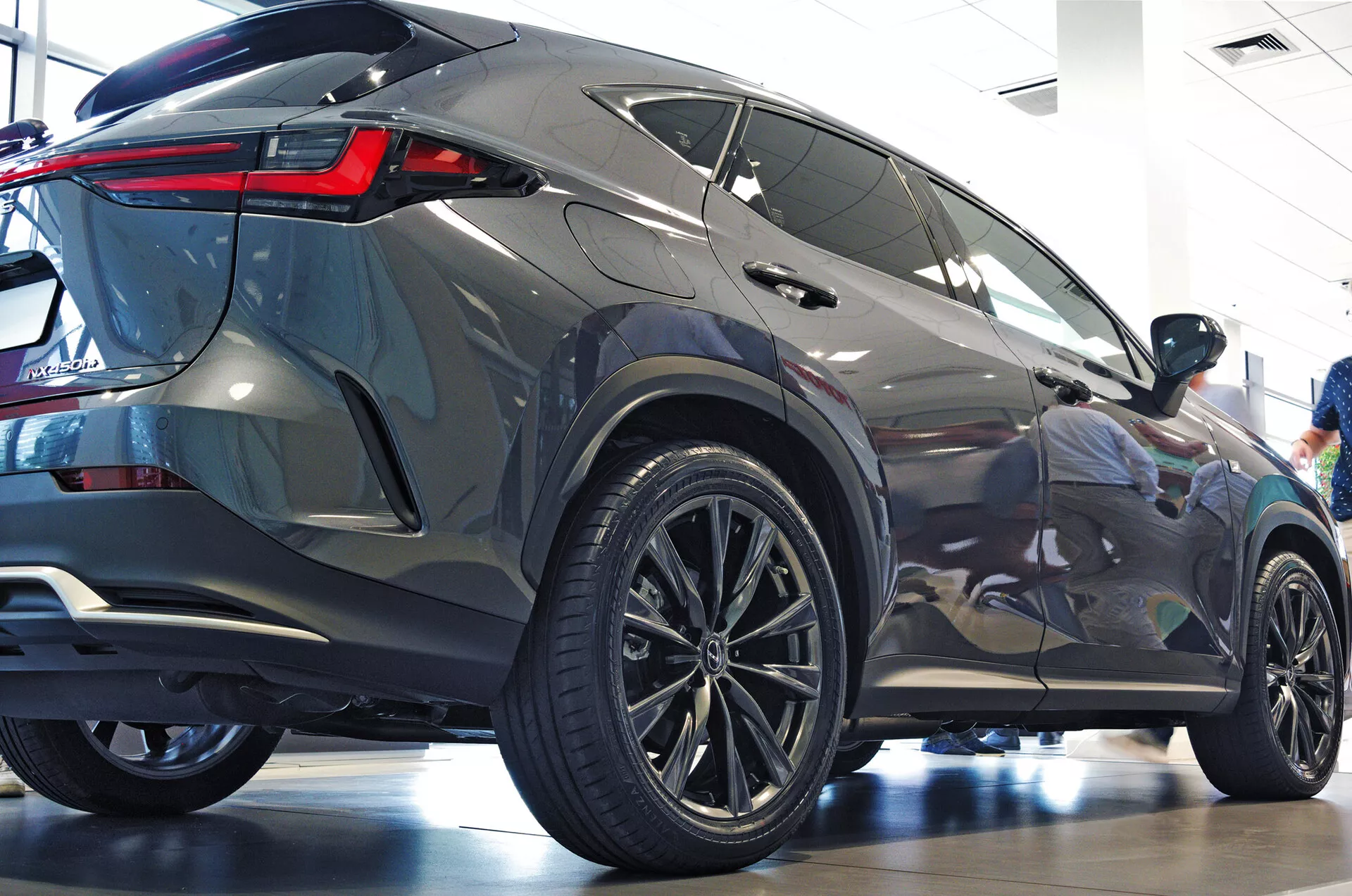 Angolo posteriore tre quarti del SUV Lexus NX 2021 che mostra il profilo laterale e posteriore con grandi ruote in lega multirazze, pneumatici a profilo ribassato, fanali posteriori LED angolari e nitidi, carrozzeria metallizzata, finestrini oscurati, spoiler posteriore integrato e ambiente da showroom pulito che enfatizza l'estetica moderna e le caratteristiche lussuose