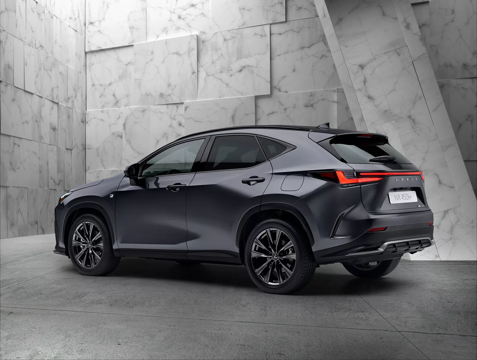 Vista posteriore a tre quarti di un moderno SUV Lexus NX 2021 ambientato in uno showroom. La vista mostra chiaramente il lato destro e posteriore del veicolo, evidenziando i fanali posteriori con design angolare a LED, la vernice scura e lucida, le ruote multi-spoke e la linea del tetto con possibili barre portatutto. Il fondo neutro e texturizzato mette in risalto il design sofisticato del SUV.