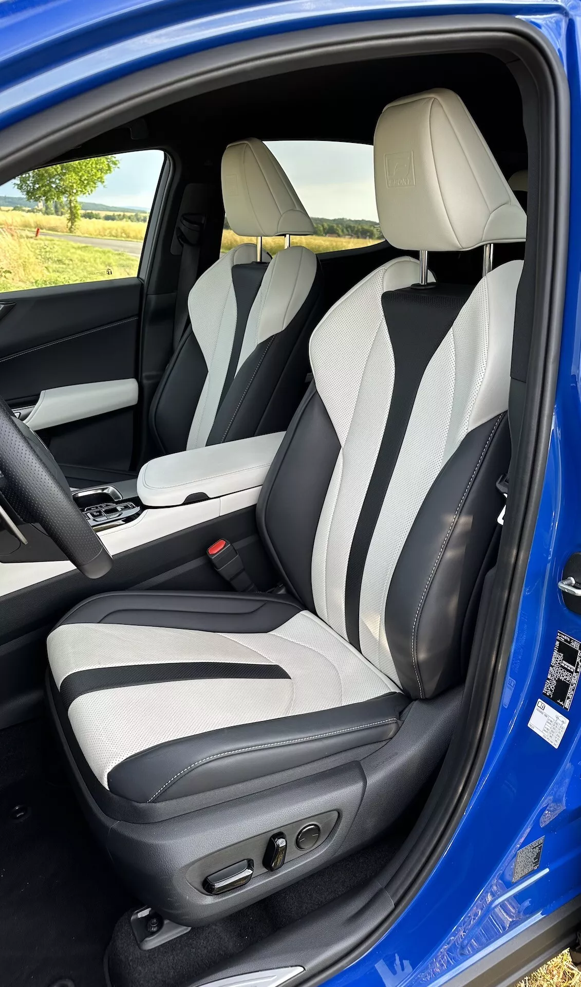 Vista interna attraverso la porta aperta del lato conducente di una Lexus NX 2021, che mostra l'area del conducente da una leggera elevazione, con tappezzeria in pelle bicolore, controlli avanzati del sedile del conducente, rinforzi laterali, poggiatesta integrati e console centrale con leva del cambio automatica