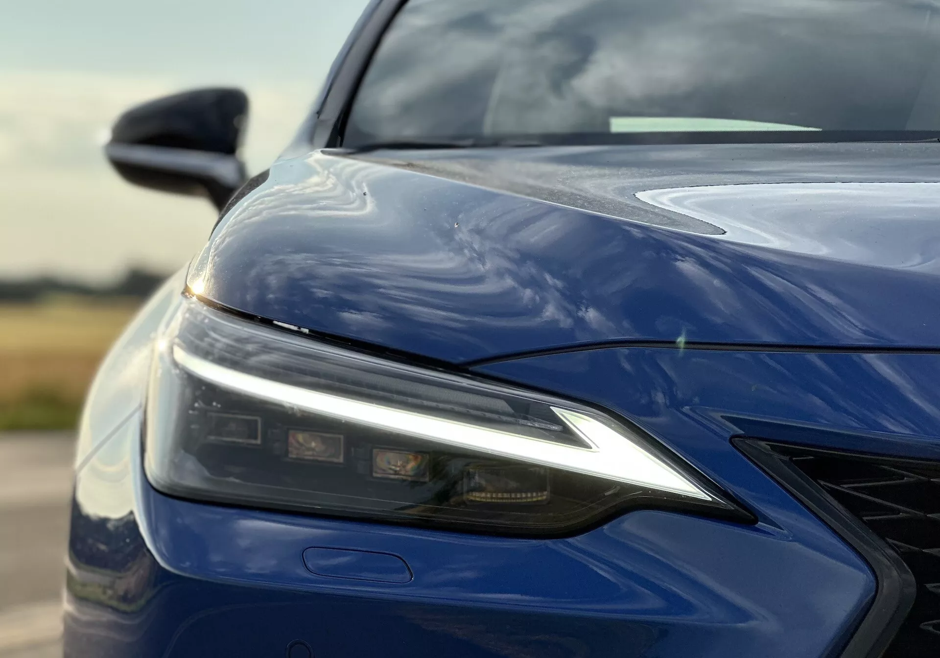 Primo piano del quarto anteriore sinistro del Lexus NX 2021 con montaggio del faro LED, cofano scolpito, paraurti anteriore con griglia e finitura blu profondo lucido, sottolineando la tecnologia di illuminazione avanzata e il design aerodinamico.