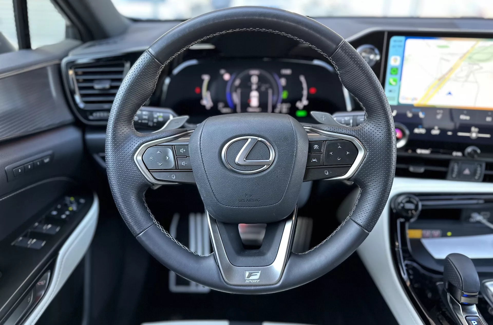 Interni della Lexus NX 2021 con focus sul volante multifunzione rivestito in pelle con emblema Lexus e logo F SPORT, quadro strumenti digitale che indica la propulsione ibrida, e scorcio del sistema di navigazione GPS sul sistema di infotainment