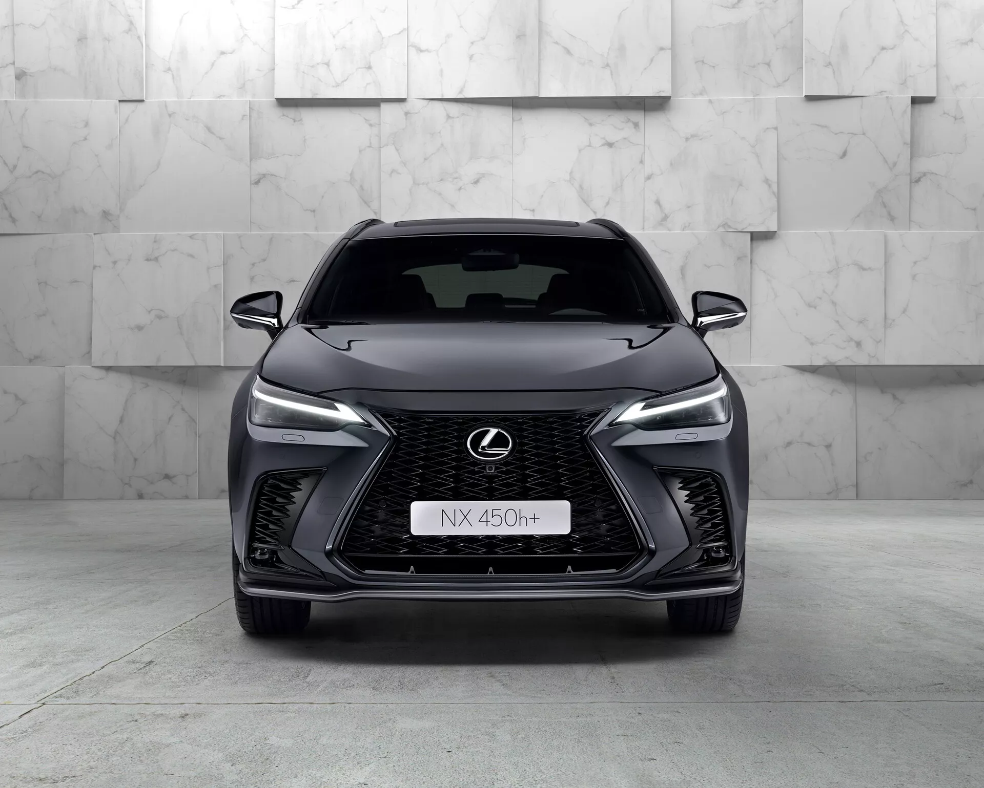 Vista frontale del Lexus NX 450h+ 2021, un moderno SUV elegante che mostra la sua prominente griglia a forma di fuso con disegno a rete, fari LED affusolati, emblema Lexus cromato con contorno blu e design aggressivo del paraurti anteriore. La natura ibrida dell'auto è indicata dal badge 'h+' sulla griglia. L'ambiente dello studio mette in risalto le curve e le caratteristiche del veicolo con un'illuminazione pulita e controllata.