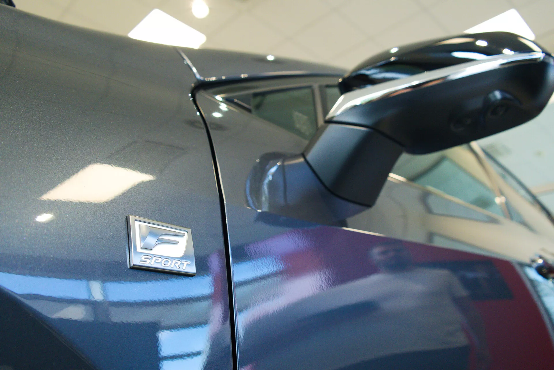 Primo piano di un'immagine di una Lexus NX 2021, con un focus sullo specchietto laterale e sul badge 'Sport'. La foto, scattata da un'angolazione bassa verso l'alto, mostra lo specchietto aerodinamico sulla carrozzeria di colore scuro. Il badge 'Sport', indicativo di una variante ad alte prestazioni, è posizionato sopra il passaruota con una ridotta profondità di campo che mette in risalto l'estetica del design.