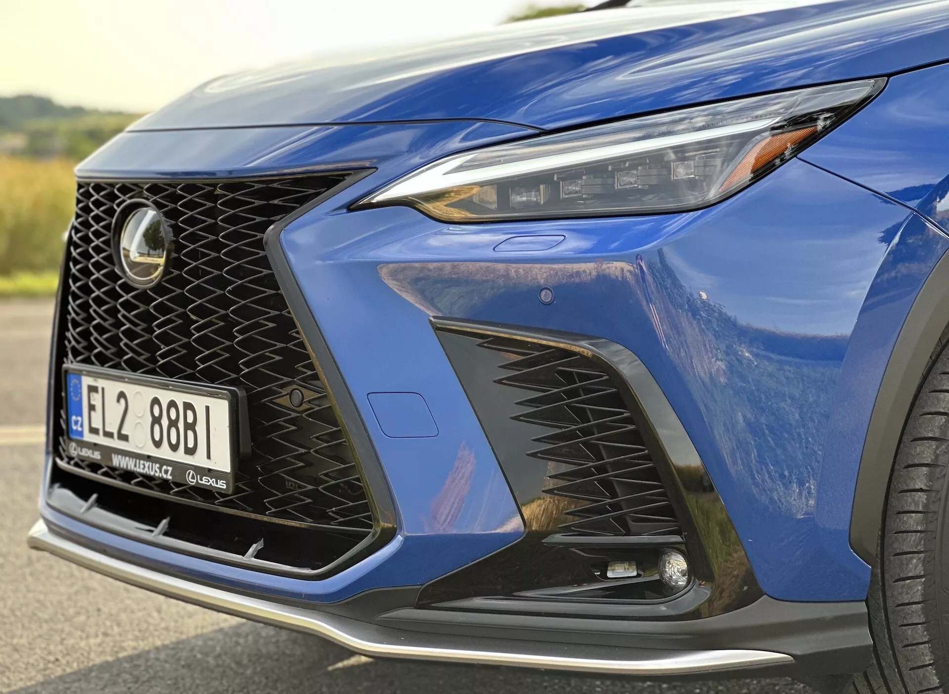 Primo piano dell'angolo anteriore del Lexus NX 2021 che mostra il faro con tecnologia LED, il design distintivo della griglia a maglia e il paraurti anteriore aerodinamico con prese d'aria, evidenziato da una vernice metallizzata blu scuro e un'occhiata a un cerchio in lega di lusso.