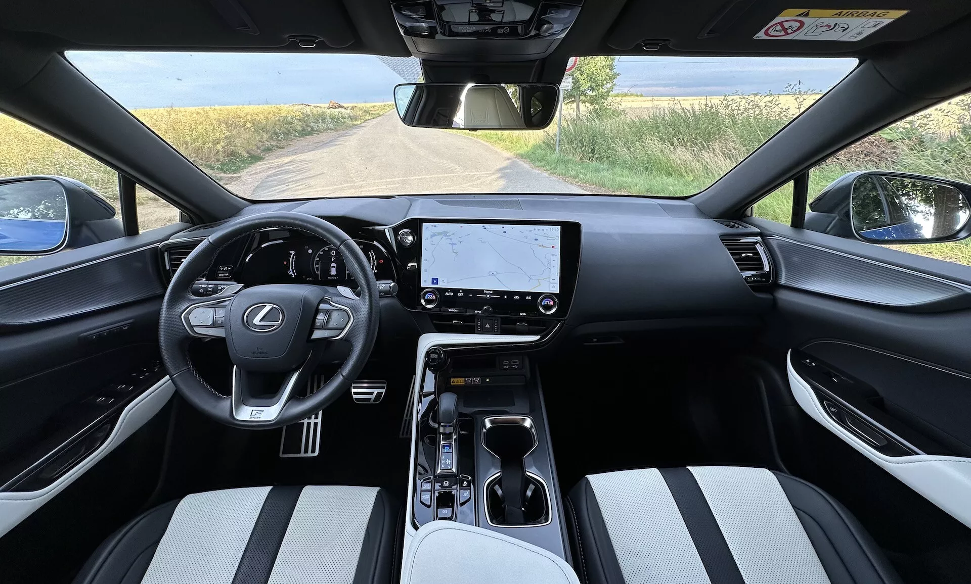 Vista interna del Lexus NX 2021 dalla prospettiva del conducente con volante centrato con logo Lexus e pulsanti di controllo, un grande touchscreen per l'infotainment sul cruscotto, controlli minimalisti per il clima, selettore di cambio automatico sulla consolle centrale con portabicchieri e vano portaoggetti, sedili rivestiti in materiale bicolore, e una vista parziale di un paesaggio rurale attraverso il parabrezza.