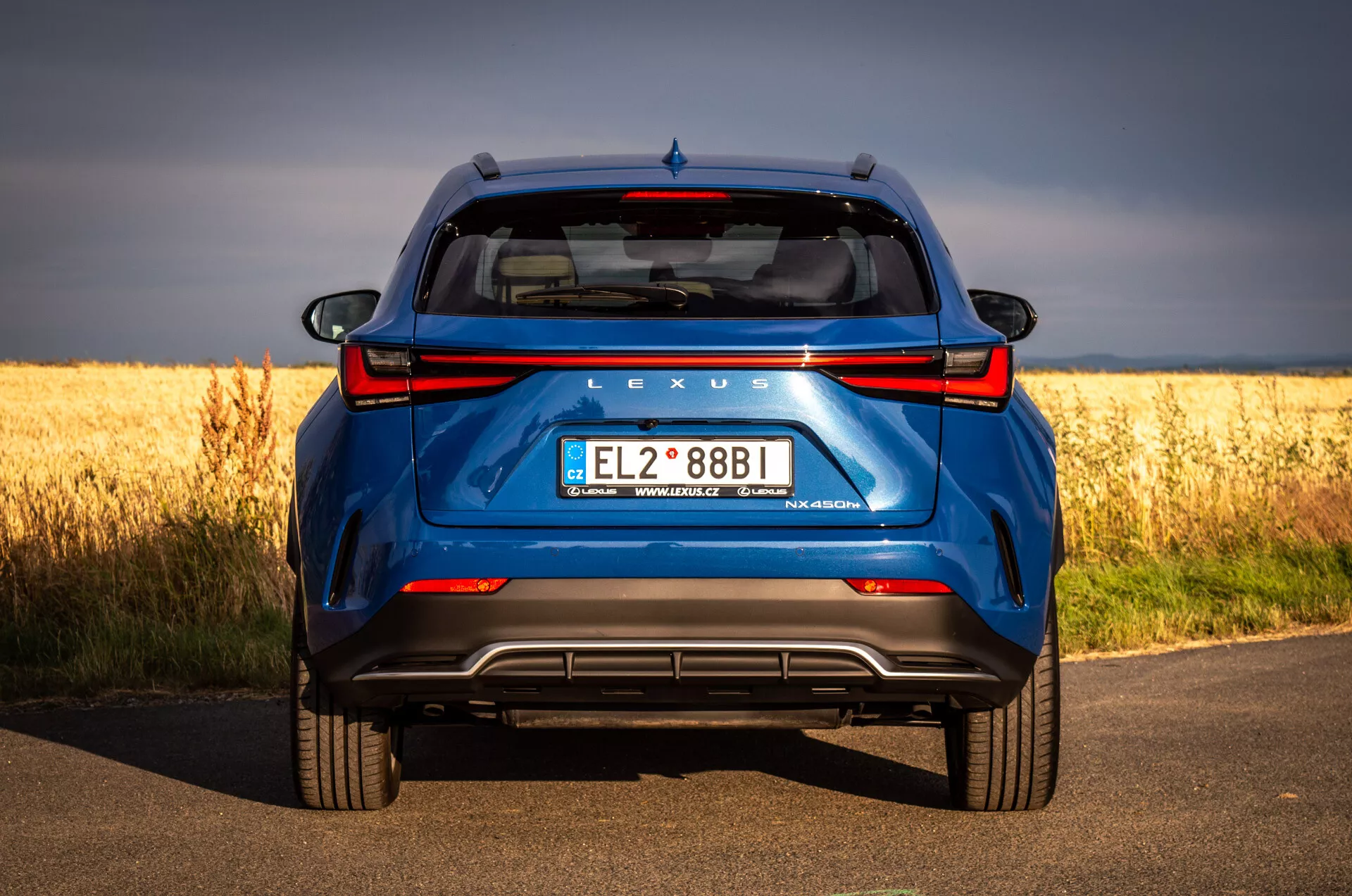 Vista posteriore della Lexus NX blu SUV 2021 su strada asfaltata con campo dorato sullo sfondo, evidenziando la tecnologia ibrida e le caratteristiche di design