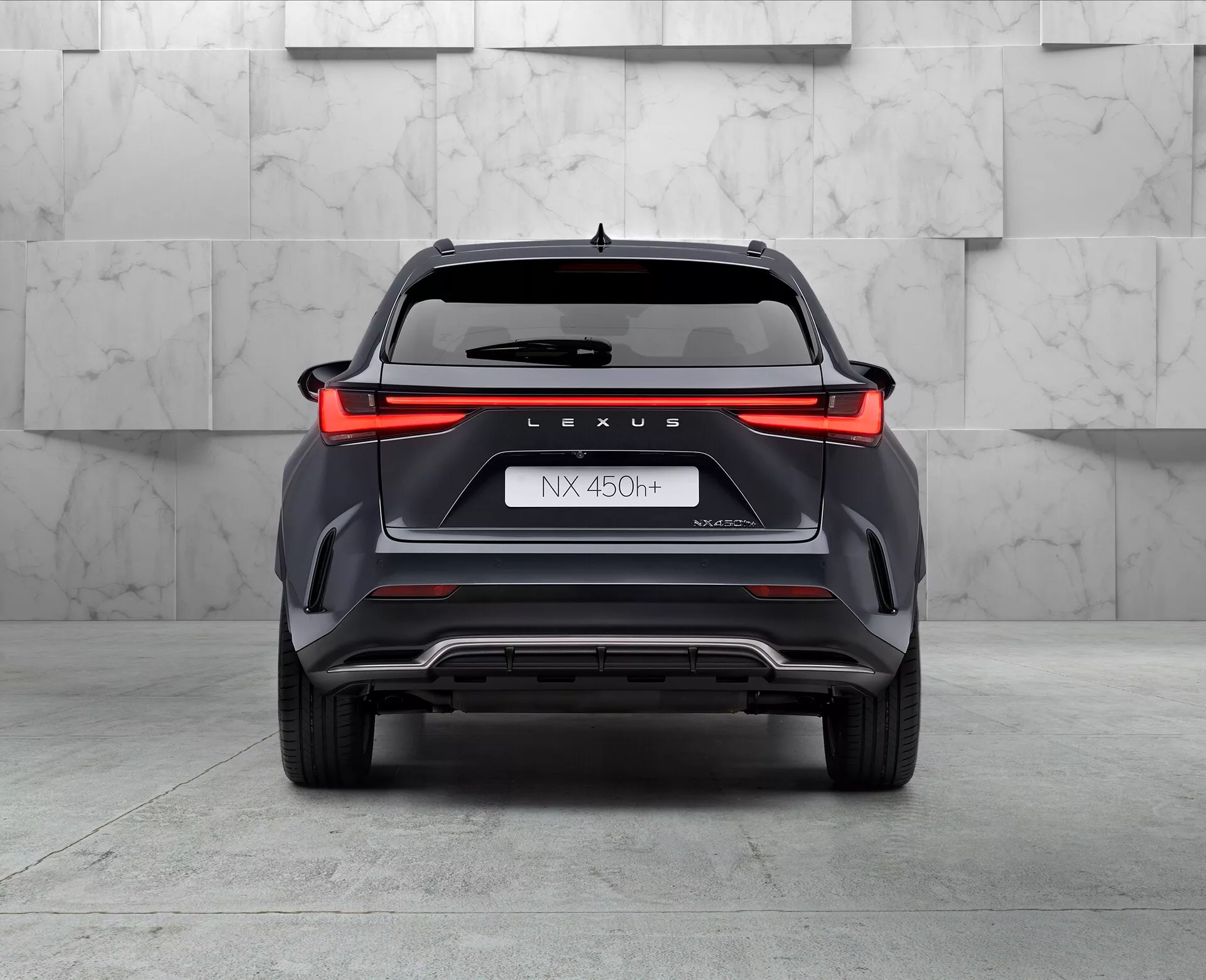 Vista posteriore del SUV ibrido Lexus NX 450h, catturata da un angolo basso leggermente inclinato a sinistra, che mostra il design della luce posteriore a tutta larghezza, il marchio 'LEXUS', la designazione del modello 'NX 450h', il nome 'NEXUS' e il simbolo dell'ibrido 'h'. Il paraurti con elementi di stile angolari, caratteristiche di sfiato e una piastra di protezione in argento, con una punta di scarico visibile sul lato sinistro, il tutto ambientato in un ambiente da showroom con pavimento lucido e sfondo neutro.