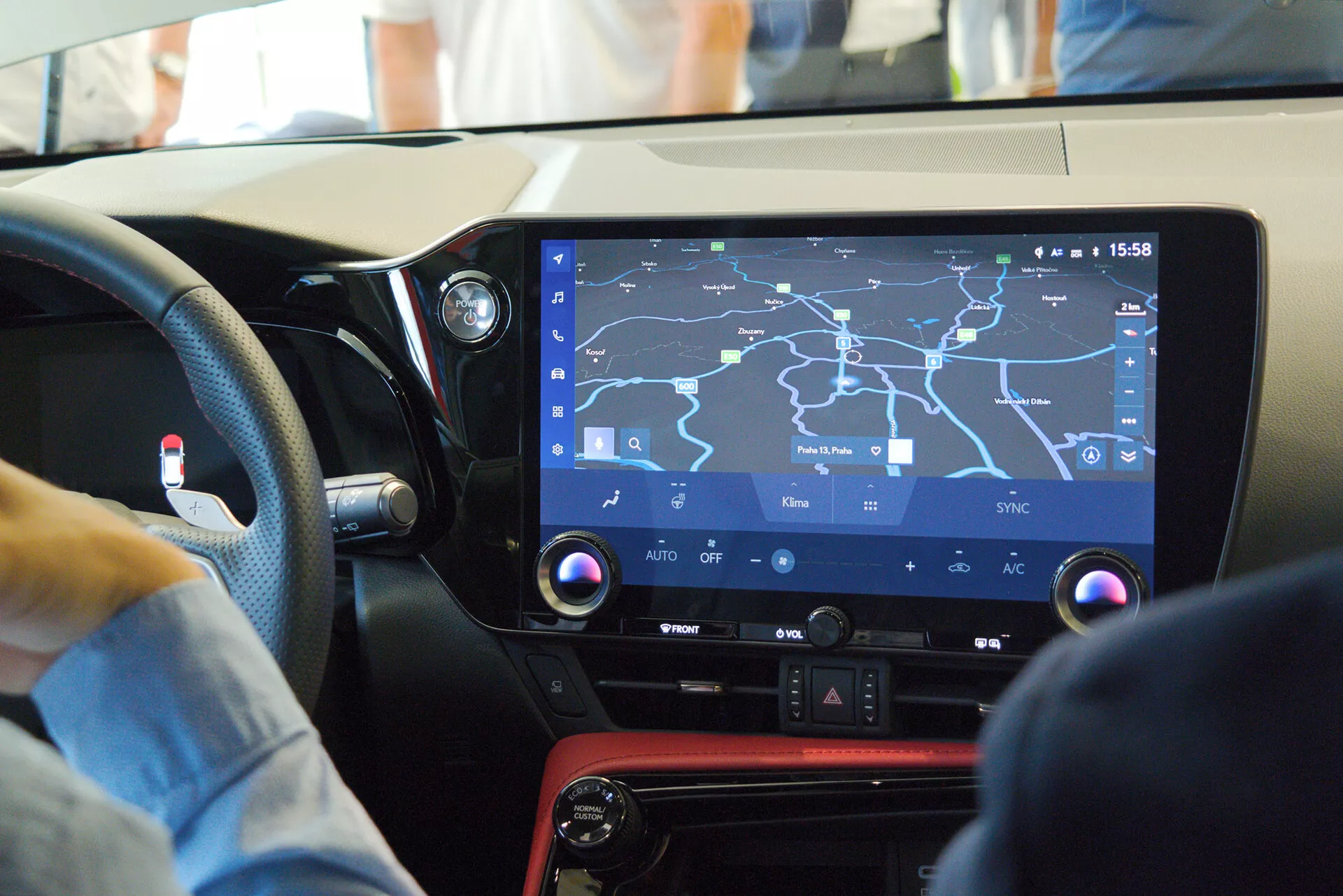 Vista interna di un'auto moderna focalizzata sulla console centrale e sul sistema di infotainment dalla prospettiva del passeggero, mostrando un grande schermo touchscreen con una mappa di navigazione, controlli fisici per le funzioni del clima, un pannello nero lucido con accento rosso e suggerendo un design orientato al guidatore in un ambiente vivace.