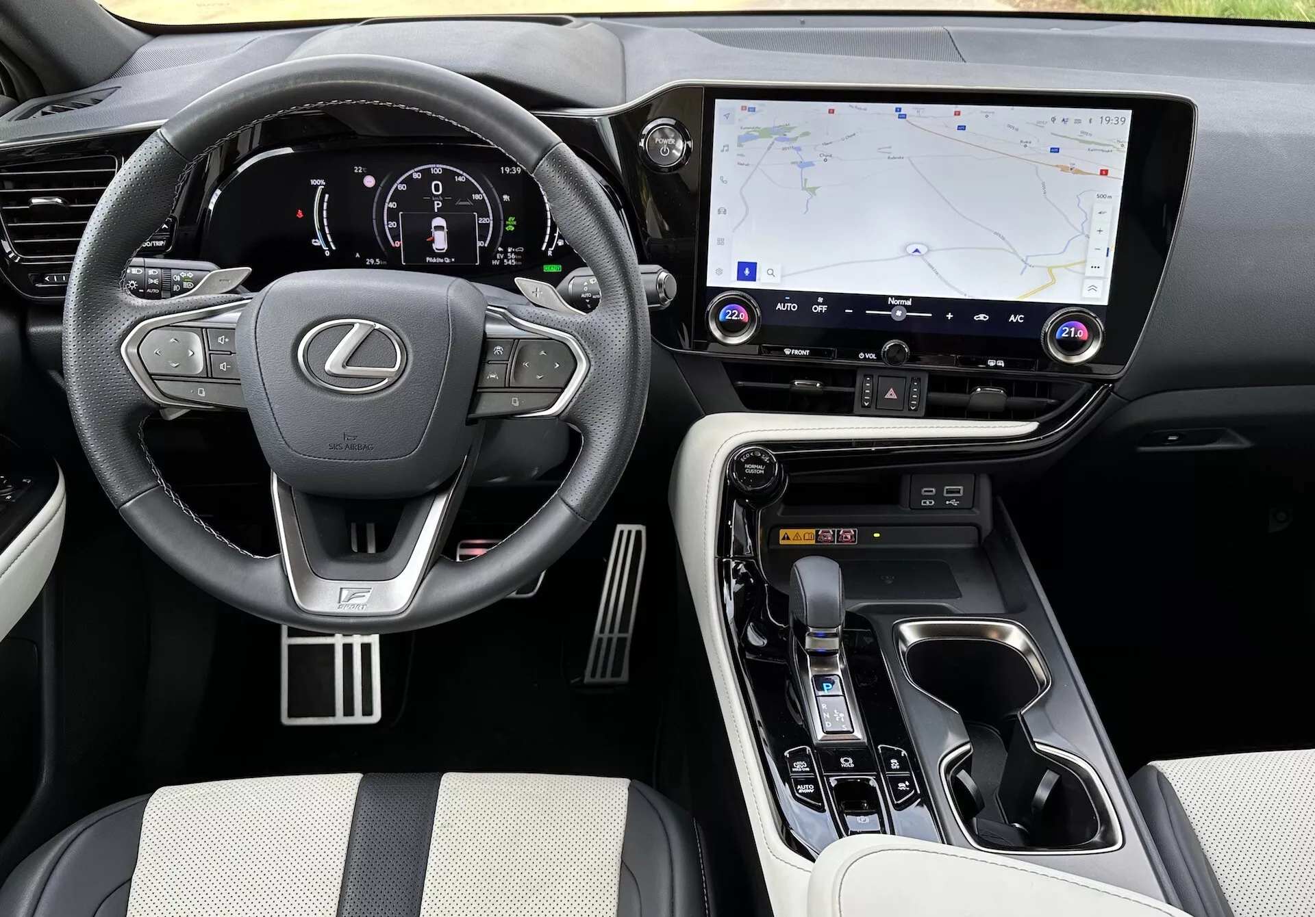Vista interna del Lexus NX 2021 che mostra l'area del guidatore e la console centrale da un angolo elevato, con il logo Lexus sul volante con comandi integrati, un cluster strumenti digitale illuminato, un grande sistema di infotainment touchscreen con navigazione GPS, pulsanti per il controllo del clima e dell'audio, un selettore del cambio ergonomico, controlli della modalità di guida, materiali di lusso sui sedili e punti di contatto, accenti metallici, e portabicchieri visibili e vano di stoccaggio.