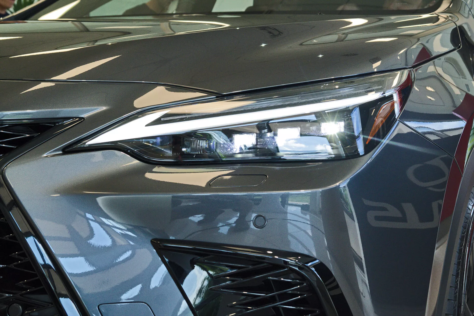 Primo piano dell'estremità anteriore di una Lexus NX 2021, in particolare del faro lato conducente con tecnologia di illuminazione avanzata a LED e parte della griglia dallo stile aggressivo. L'immagine cattura il luccichio riflettente della vernice argentata metallizzata e le linee dinamiche che contribuiscono all'estetica aerodinamica del veicolo. Fotografato all'interno, probabilmente in uno showroom, evidenziando il design lussuoso e la finitura di alta qualità del veicolo.