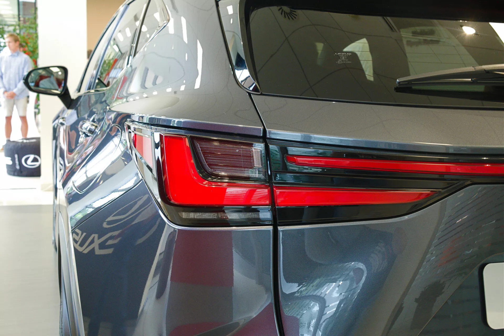 Primo piano del 2021 Lexus NX da un angolo posteriore di tre quarti che evidenzia il design elegante del fanale posteriore a LED, parte del pannello posteriore e una porzione del coperchio del bagagliaio. L'immagine mostra la vernice scura e lucida dell'auto, le linee aerodinamiche della carrozzeria e suggerisce un'ambientazione interna di showroom con riflessi di un altro veicolo, elementi architettonici e una persona che cammina sullo sfondo.