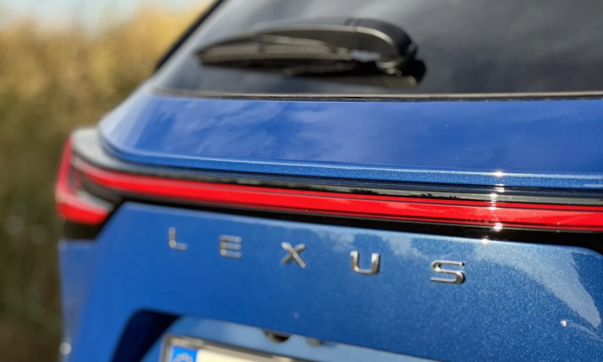 Vista ravvicinata dell'area di branding posteriore della Lexus NX 2021 con distintivo Lexus argento, una striscia di luce rossa sopra il nome, su un corpo blu metallico in condizioni di luce diurna