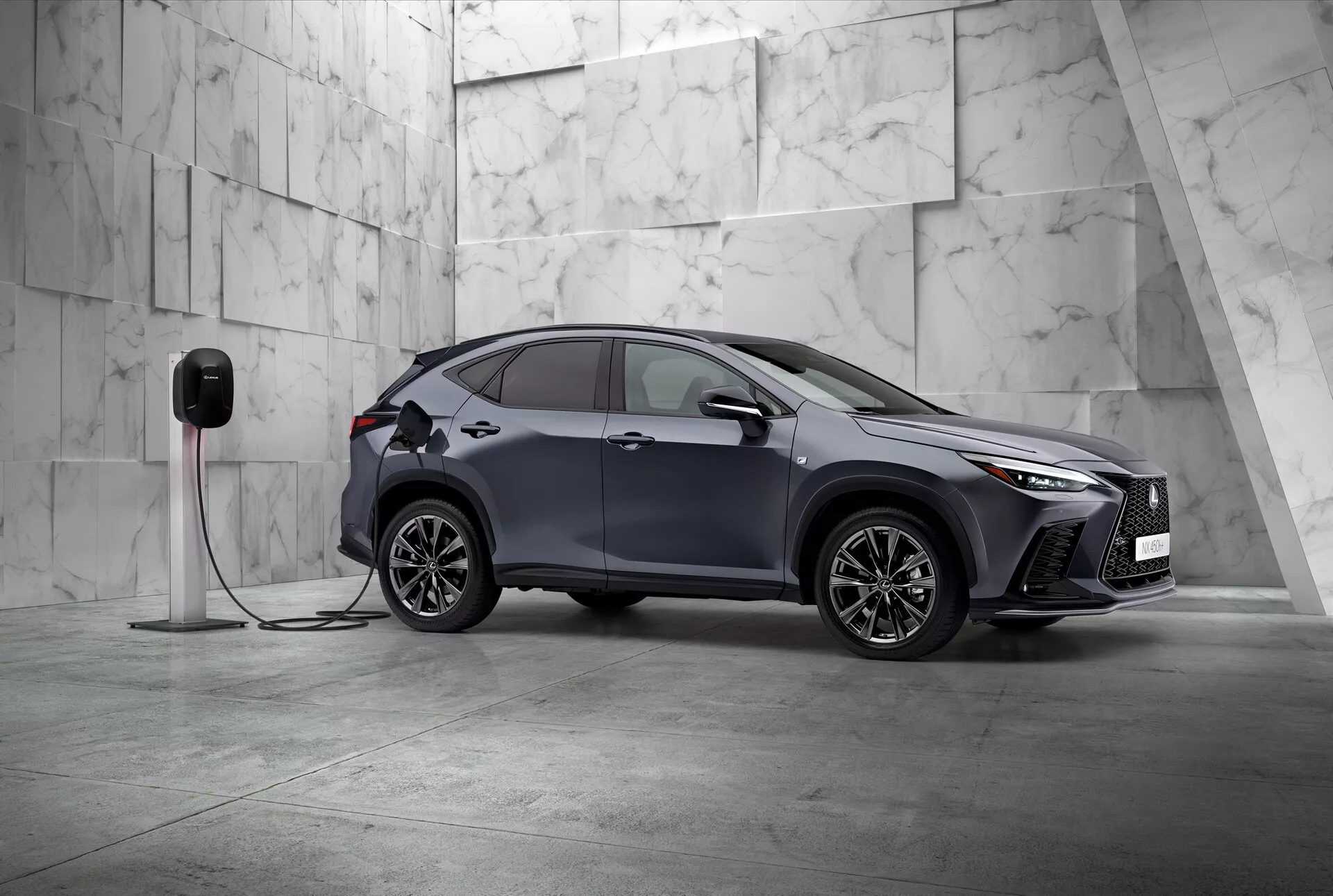 Questa immagine mostra il Lexus NX 2021, un moderno SUV compatto con un design elegante e sportivo, fotografato da una vista anteriore di tre quarti. Sono visibili il lato sinistro del veicolo e parzialmente il fronte, posizionati in diagonale in un ambiente minimalista adornato con grandi piastrelle di marmo chiaro, suggerendo un'atmosfera premium. Il Lexus NX vanta fari LED angolari distintivi e una griglia anteriore prominente, enfatizzando la sua natura sofisticata. Si notano specchietti laterali aerodinamici, cerchi in lega intricati con più raggi e maniglie delle porte incassate che evidenziano il design sportivo e avanzato. Un dettaglio cruciale è la connessione dell'auto a una stazione di ricarica elettrica, indicando che si tratta di un veicolo elettrico o ibrido plug-in. Il parafango anteriore include una porta di ricarica, adatta alle sue capacità elettriche. Questa immagine statica si concentra sulla presentazione del design del veicolo e delle sue caratteristiche tecnologiche in una composizione equilibrata e tranquilla con ampio spazio negativo, esemplificando esclusività e innovazione nel design automobilistico.