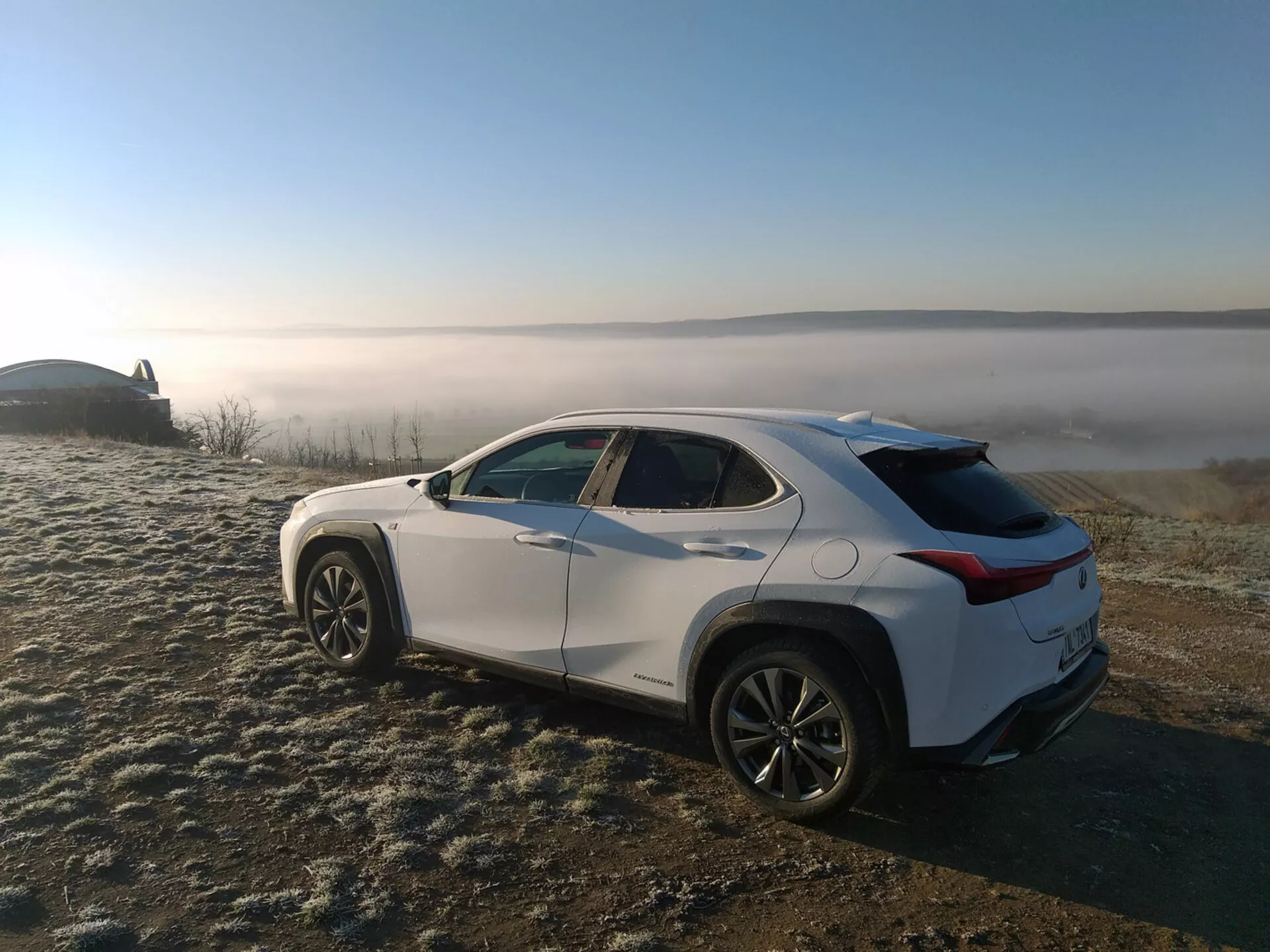 SUV crossover Lexus UX del 2019 parcheggiato su erba coperta di brina con sfondo di paesaggio nebbioso, mettendo in mostra il profilo del veicolo da un angolo posteriore di tre quarti che evidenzia le linee caratteristiche laterali, gli archi delle ruote, la linea del tetto che si assottiglia e i cerchi in lega.