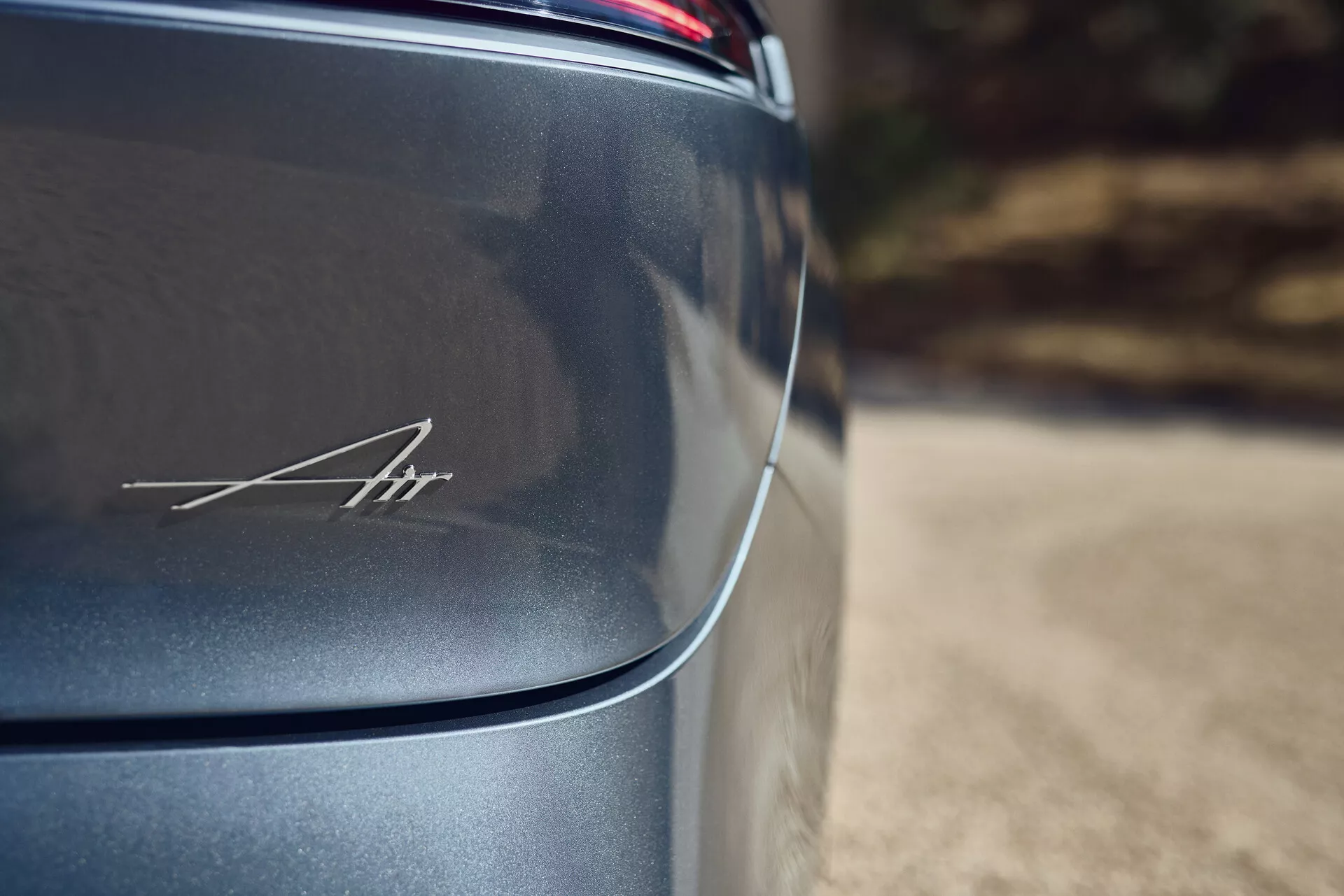 Vista ravvicinata e angolata del pannello posteriore sul lato del conducente del Lucid Air 2022 di prima generazione, che illustra il suo design elegante e aerodinamico. La finitura metallica scura, probabilmente in tonalità di grigio o nero, brilla sotto una luce naturale morbida. Il distintivo elegante 'Air' in corsivo nella sezione inferiore destra è messo in evidenza attraverso una profondità di campo ridotta e un effetto bokeh, mettendo in risalto il carattere lussuoso ed esclusivo del veicolo.