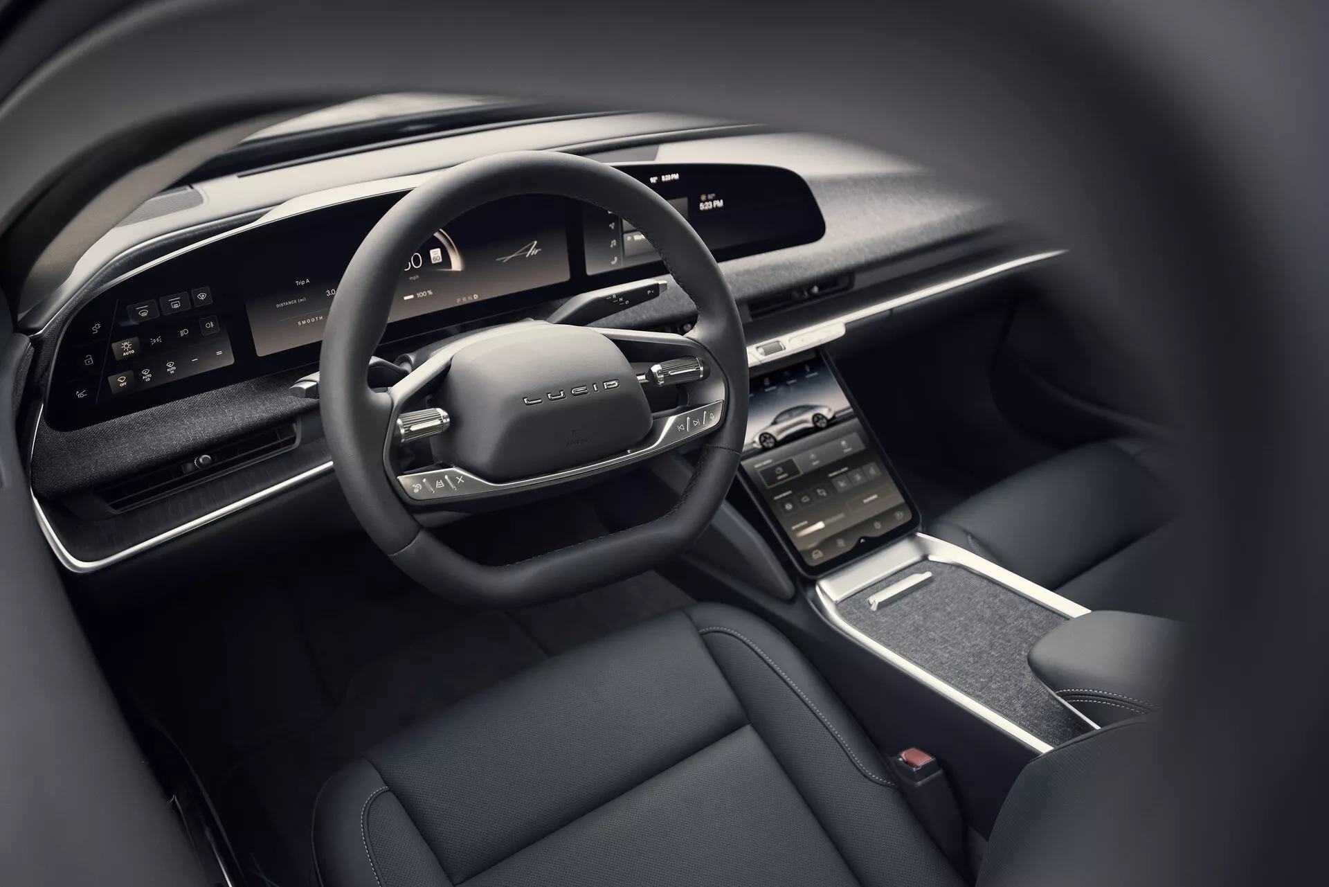 Vista ravvicinata dell'abitacolo del guidatore della Lucid Air 2022 dalla prospettiva del sedile posteriore, che mostra il volante multifunzione moderno con comandi, l'ampio cruscotto digitale, il touchscreen centrale orientato verticalmente con prese d'aria circostanti, il console centrale elegante con pochi pulsanti fisici e un interno di lusso con schema di colori scuri caratterizzato da materiali di alta qualità.