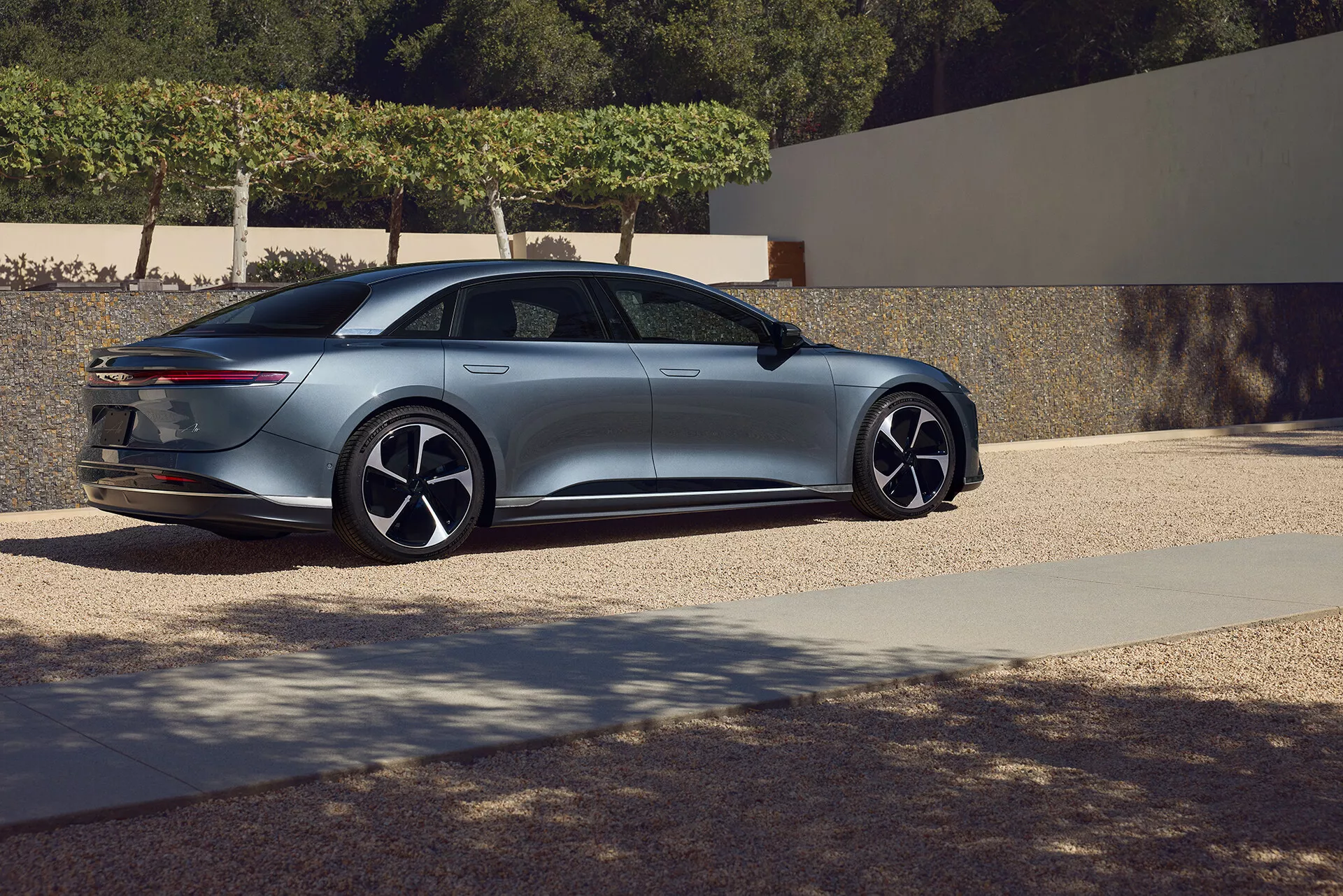 Vista laterale posteriore a tre quarti della berlina Lucid Air 2022 di prima generazione. L'inquadratura a bassa angolazione enfatizza il design elegante e aerodinamico del veicolo, caratterizzato da una linea del tetto fluida che si integra in un cofano posteriore in stile fastback con fanali posteriori sottili e orientati orizzontalmente. Il profilo laterale mostra grandi ruote in lega multi-razza e maniglie delle porte a filo. I sottili specchietti laterali e il cofano inclinato suggeriscono caratteristiche tecnologiche avanzate, arricchendo la sensazione di lusso e modernità dell'auto. L'ambientazione sembra essere un'area residenziale, che esalta l'estetica premium.