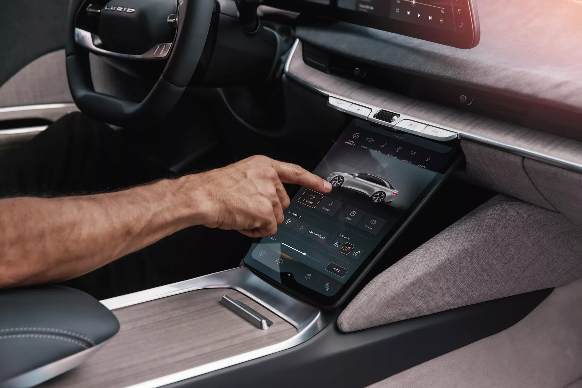 Vista dettagliata della console centrale della prima generazione di Lucid Air 2022 da un'angolazione obliqua, evidenziando un display touchscreen verticalmente orientato interagito con la mano destra di una persona, illustrando il sistema di infotainment avanzato con vari controlli funzionali e una combinazione di pannellatura tessile e metallica, suggerendo un design degli interni moderno e di lusso incentrato sulla tecnologia avanzata.