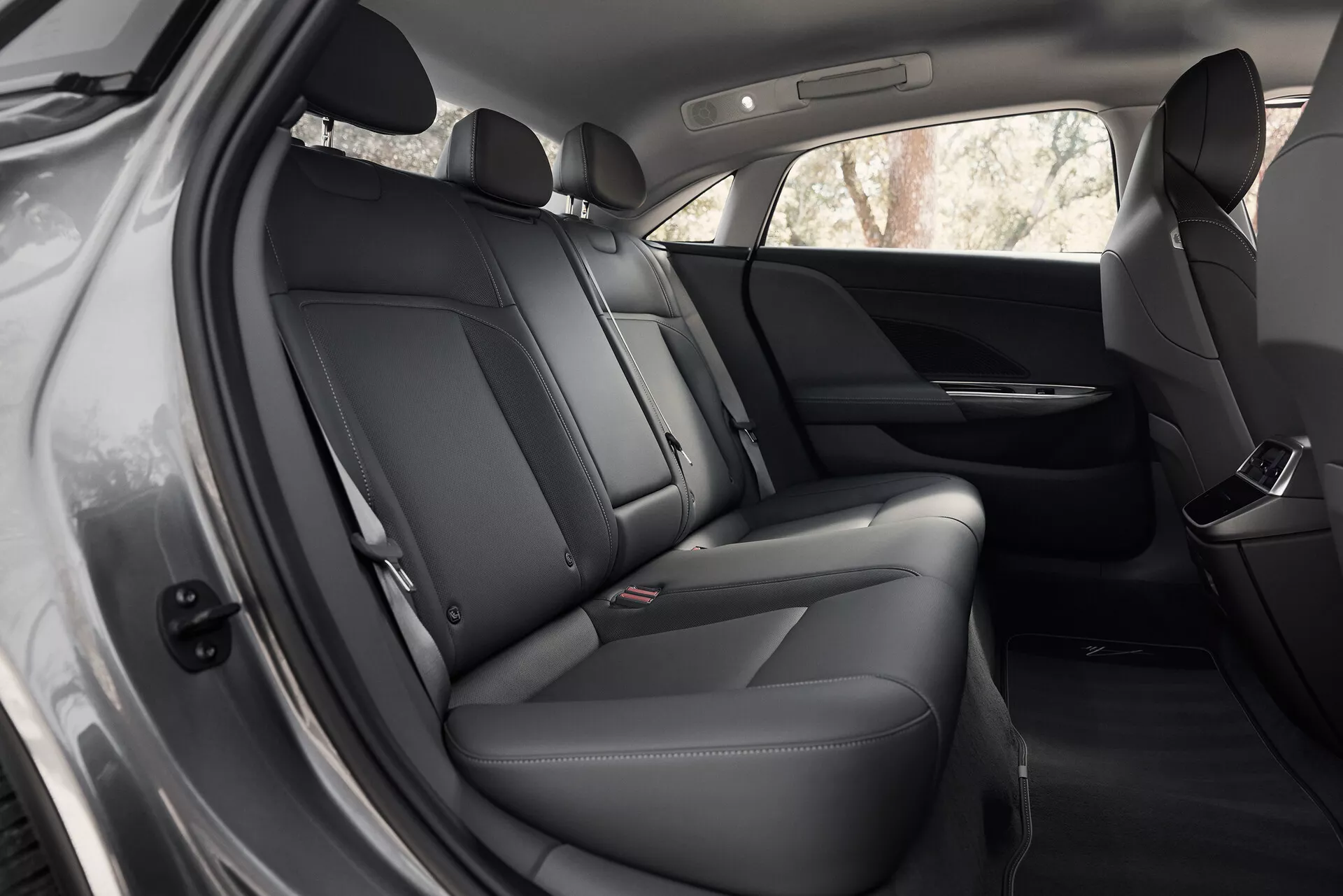 Vista interna di una Lucid Air 2022 di prima generazione, focalizzandosi sull'area dei sedili posteriori da un angolo leggermente rivolto in avanti. I sedili presentano rivestimenti in pelle scura o materiale sintetico premium con cuciture visibili. Il pannello della porta mostra un altoparlante, controllo dei finestrini elettrici e possibili vani portaoggetti. Gli schienali dei sedili anteriori e l'abitacolo sono puliti e ben mantenuti, illuminati dalla luce naturale.