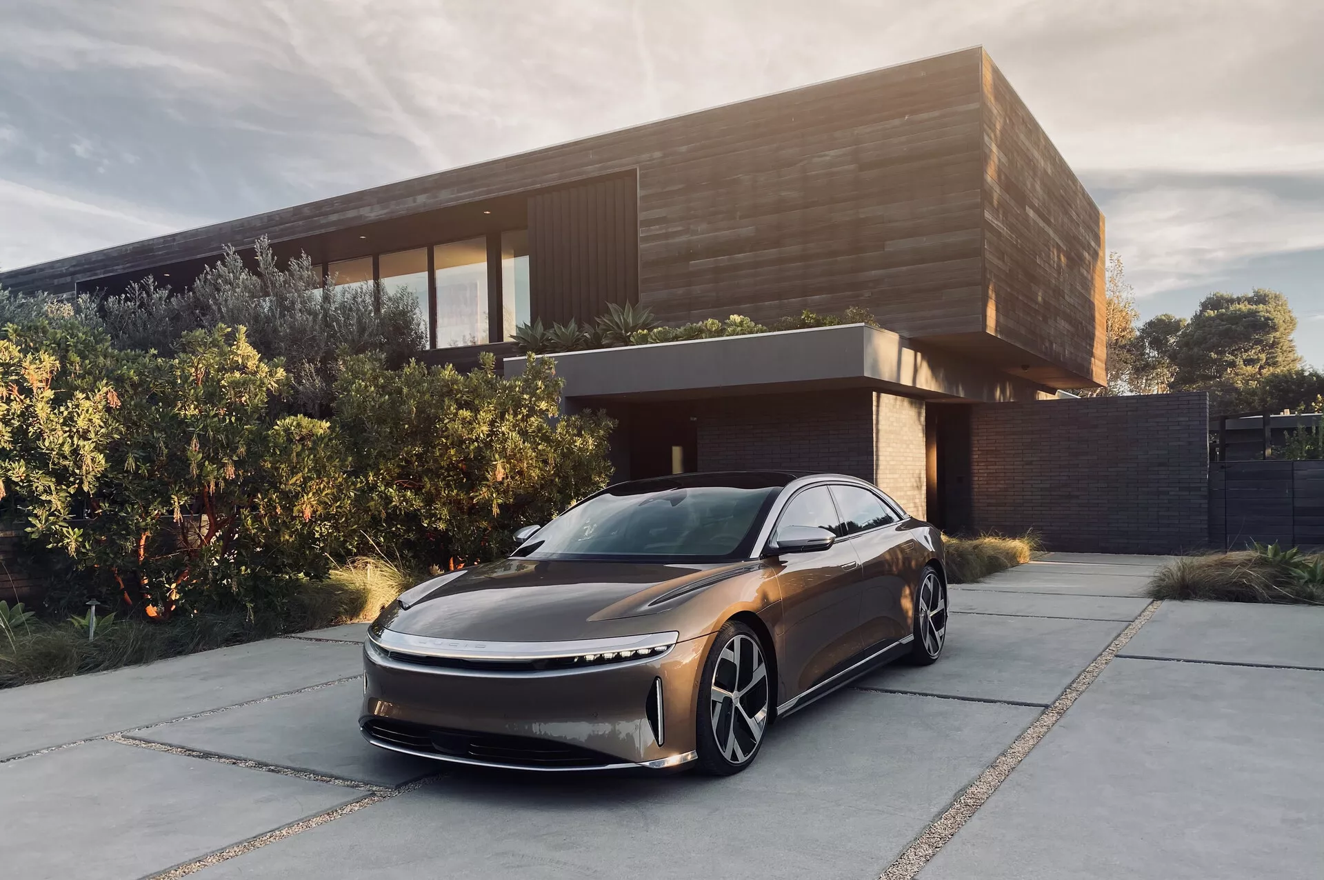 Immagine della Lucid Air 2022, una berlina moderna, parcheggiata davanti a una casa contemporanea. Ripresa da un angolo basso e frontale a tre quarti, che mostra il suo design aerodinamico ed elegante con cofano allungato e griglia sottile. Caratteristiche evidenti includono i fari a LED sottili, le grandi ruote distintive e l'assenza di specchietti laterali suggerendo l'uso di sistemi a telecamera per la visione posteriore. Il colore scuro metallizzato e l'ambientazione sofisticata esaltano la sua sensazione di lusso, suggerendo che è un veicolo elettrico destinato a un mercato facoltoso.