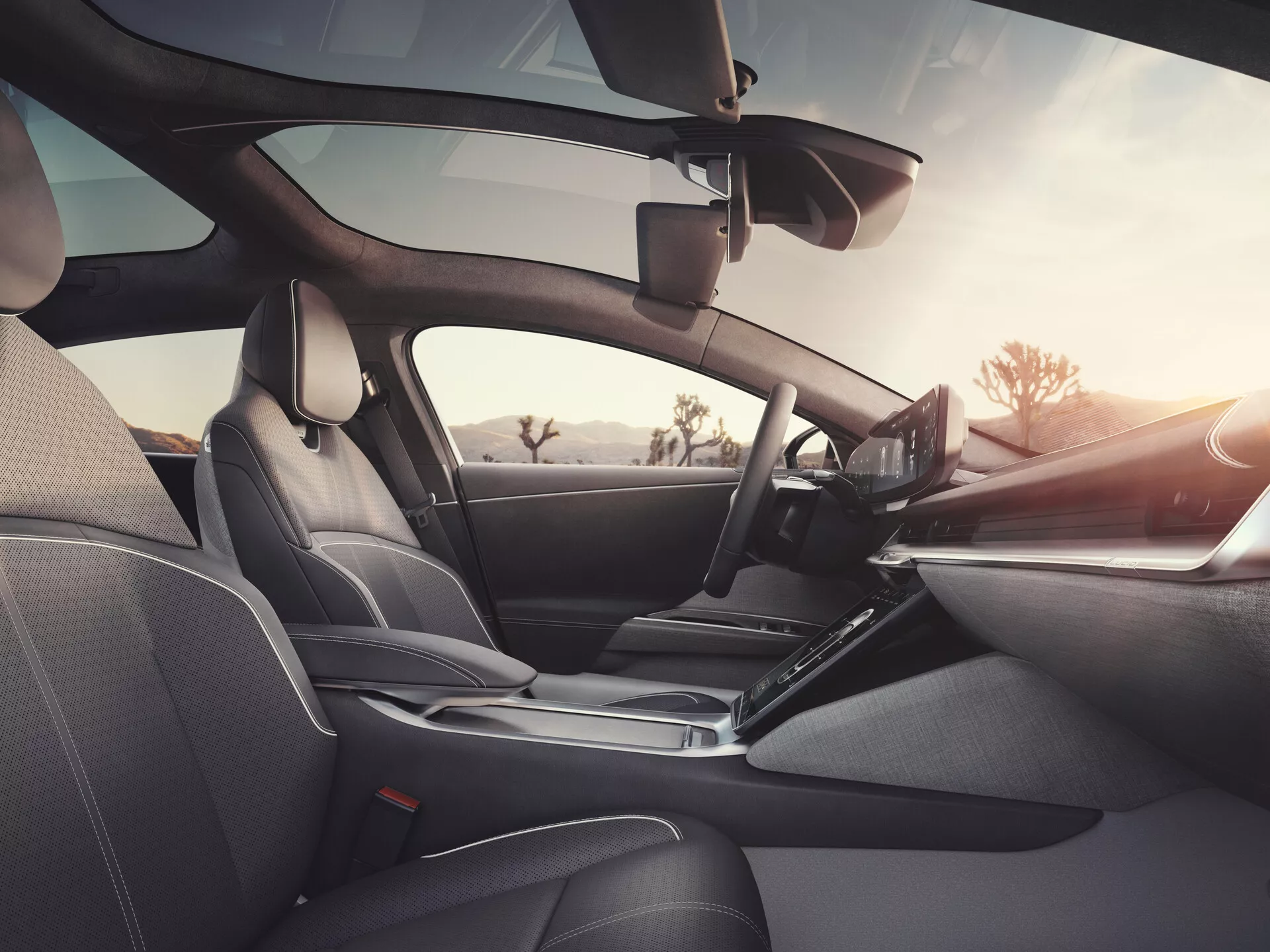 Vista interna del Lucid Air 2022 (1ª generazione) dalla prospettiva del passeggero posteriore, che mostra sedili in pelle scura, cuciture dettagliate, cruscotto elegante con volante a sinistra con comandi integrati, display di infotainment touchscreen prominente, leva del cambio ergonomica, pannelli delle portiere con comandi elettronici e altoparlanti, specchietto retrovisore visibile e possibile dispositivo di assistenza alla guida vicino al parabrezza, sottolineando il design lussuoso, tecnologia avanzata e comfort.