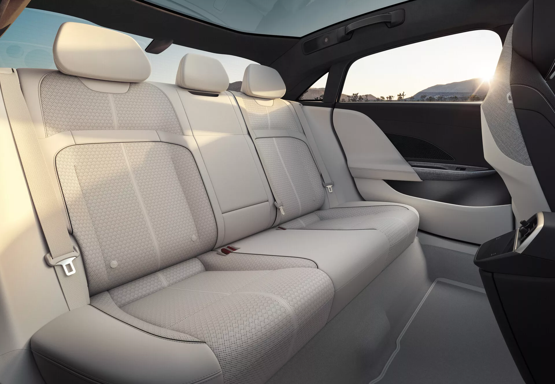 Vista dell'interno della Lucid Air 2022, focalizzata sui sedili posteriori. I sedili sono rivestiti in pelle chiara, offrendo una sensazione di ariosità e spazio. La panca posteriore ospita tre passeggeri con poggiatesta abbinati e cinture di sicurezza visibili. I pannelli delle porte si fondono perfettamente con il materiale e il design dei sedili, presentando griglie degli altoparlanti integrate. Una linea di finestrini alta e un ampio tetto panoramico oscurato migliorano il senso di privacy e protezione. Il rivestimento interno mostra un mix di tappezzeria chiara ed elementi più scuri, probabilmente di materiali di alta qualità.