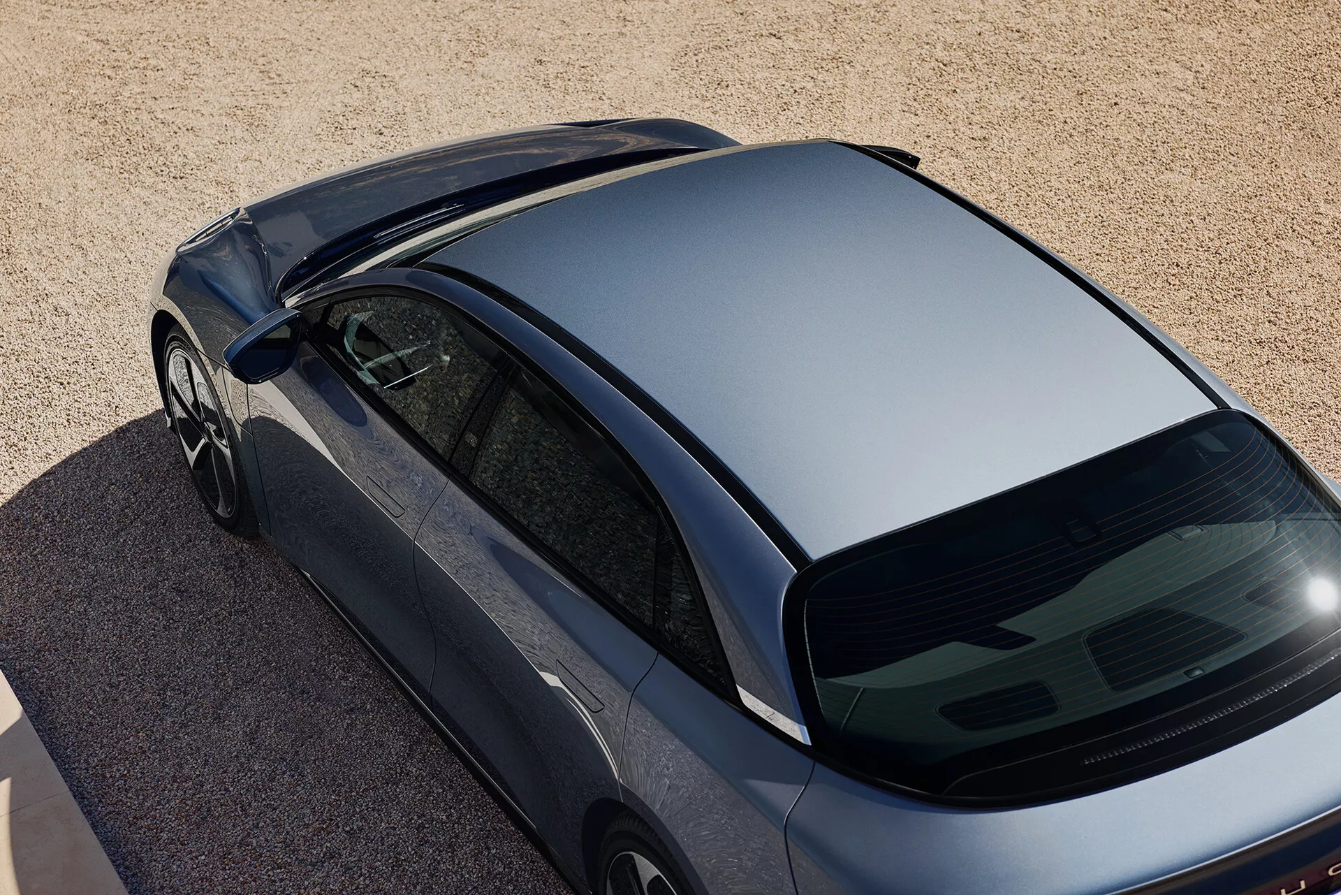 Vista aerea angolata di un'auto Lucid Air 2022 di prima generazione in colore metallico scuro, parcheggiata su una superficie di piccoli ciottoli o sabbia grossolana. La ripresa cattura la vista dall'alto, focalizzata sul tetto aerodinamico, il lunotto posteriore ripido e una parte del bagagliaio. Le caratteristiche includono finestrini senza cornice, specchietti laterali aerodinamici e l'assenza di accessori sul tetto e tergilunotto, suggerendo un design aerodinamico e moderno.