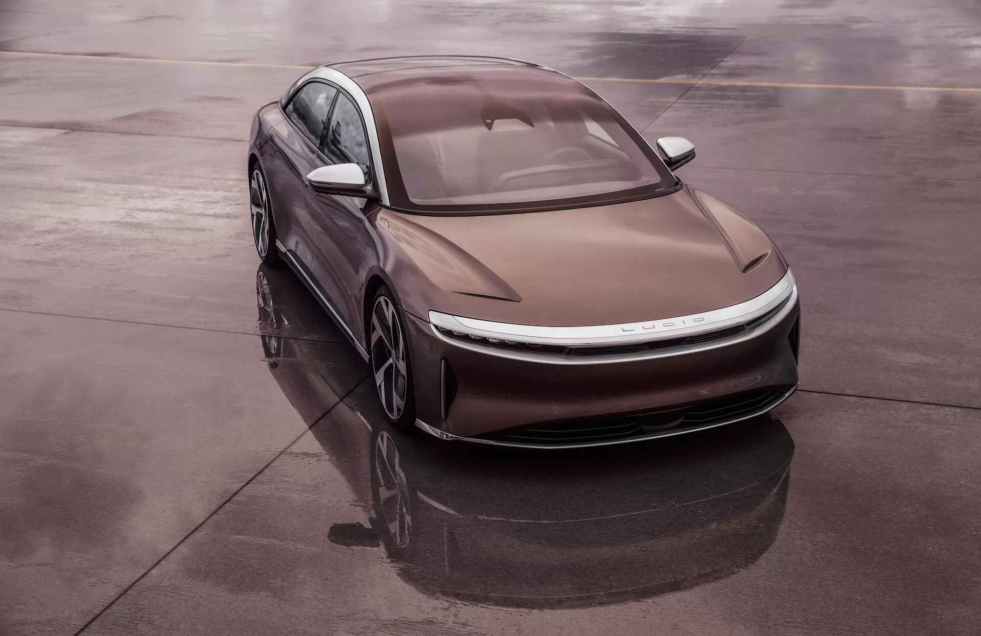 Foto dall’alto di una berlina moderna Lucid Air 2022 di prima generazione, posizionata in diagonale su una superficie riflettente come l’asfalto bagnato, che offre una vista completa del quarto anteriore sinistro del veicolo. Il design aerodinamico ed elegante presenta curve morbide, fari LED sottili e un cofano sagomato con linee marcate. La linea del tetto tipo fastback e il vetro a filo migliorano l’efficienza aerodinamica. La natura elettrica dell'auto è suggerita dall’assenza di una griglia tradizionale, un sistema di presa d’aria integrato e ruote aerodinamiche leggere.
