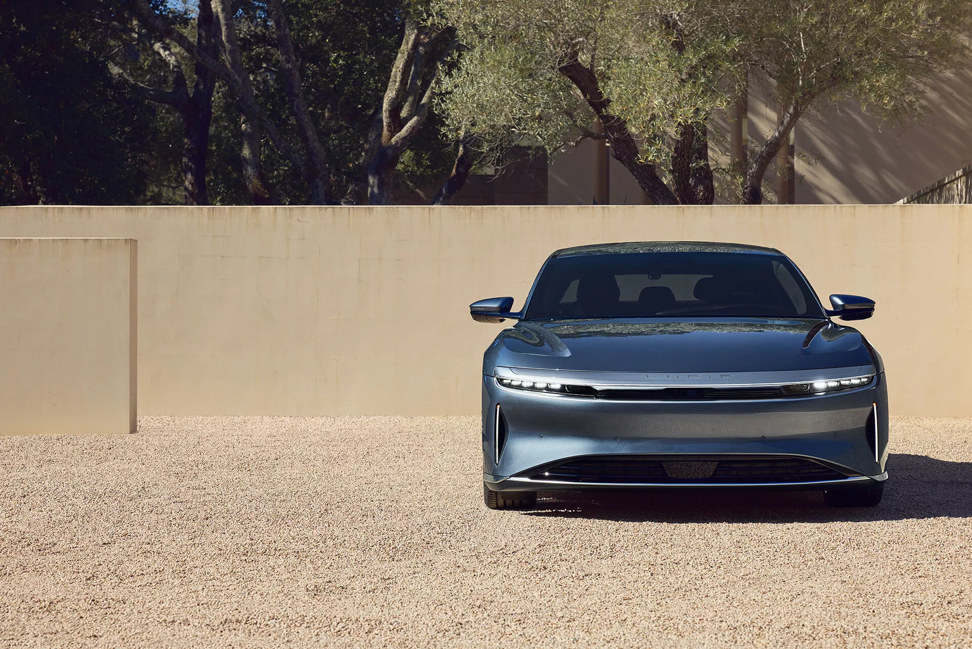 Inquadratura ravvicinata del frontale di una Lucid Air 2022 di prima generazione, evidenziando la griglia chiusa elegante, i fari LED orizzontali, il paraurti anteriore integrato, il cofano scolpito con una linea di contorno e la finitura della vernice metallizzata. L'auto è posizionata centralmente su una superficie di ghiaia sotto un'illuminazione brillante, suggerendo un'aerodinamica avanzata e un design lussuoso e moderno.