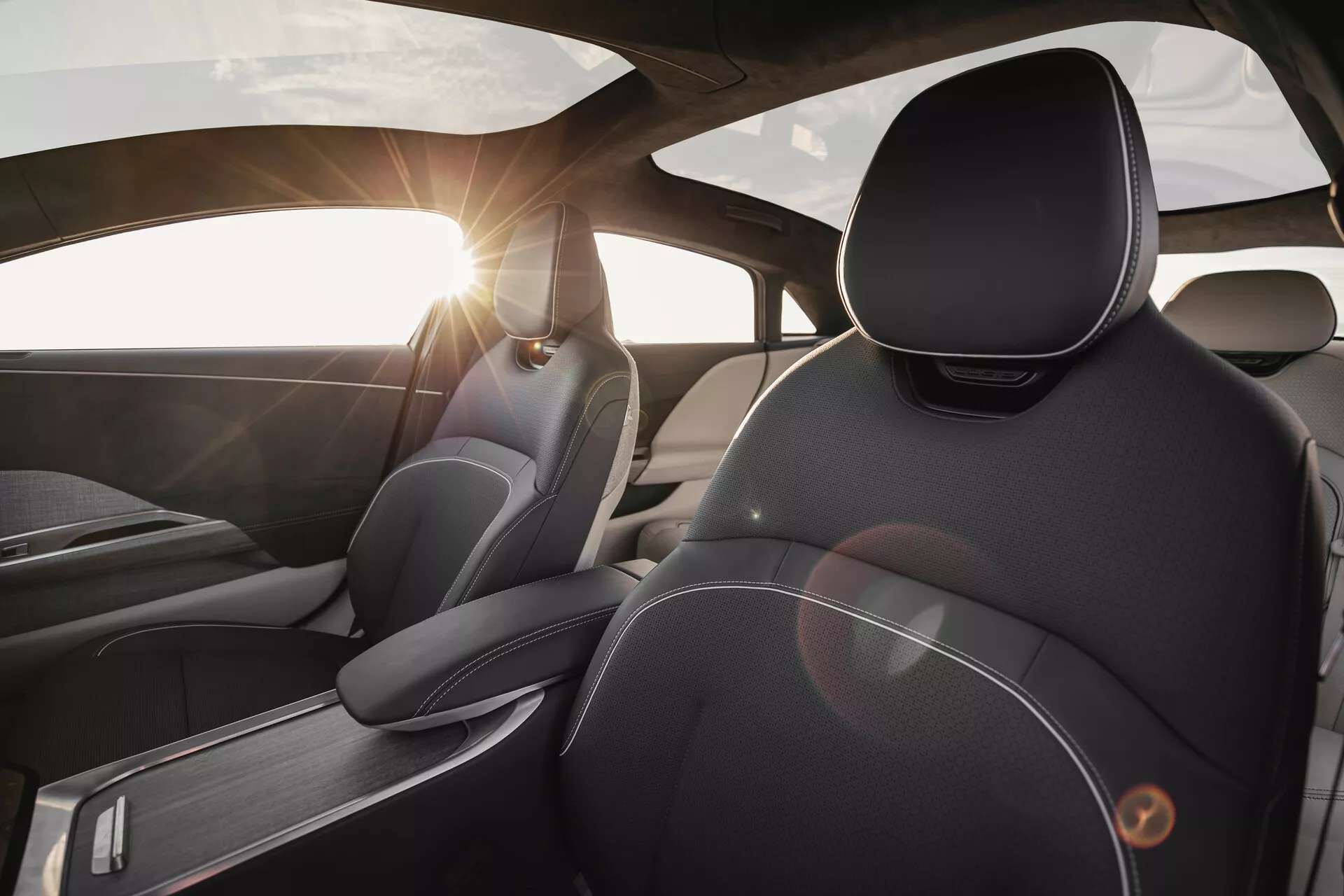 Vista interna della Lucid Air 2022 dalla prospettiva del sedile posteriore, centrata sul sedile del conducente. Si concentra su materiali e design di alta qualità, con cuciture dettagliate sui rivestimenti in pelle, poggiatesta ergonomici, accenti in metallo spazzolato e un effetto flare causato dalla luce del sole attraverso il parabrezza.