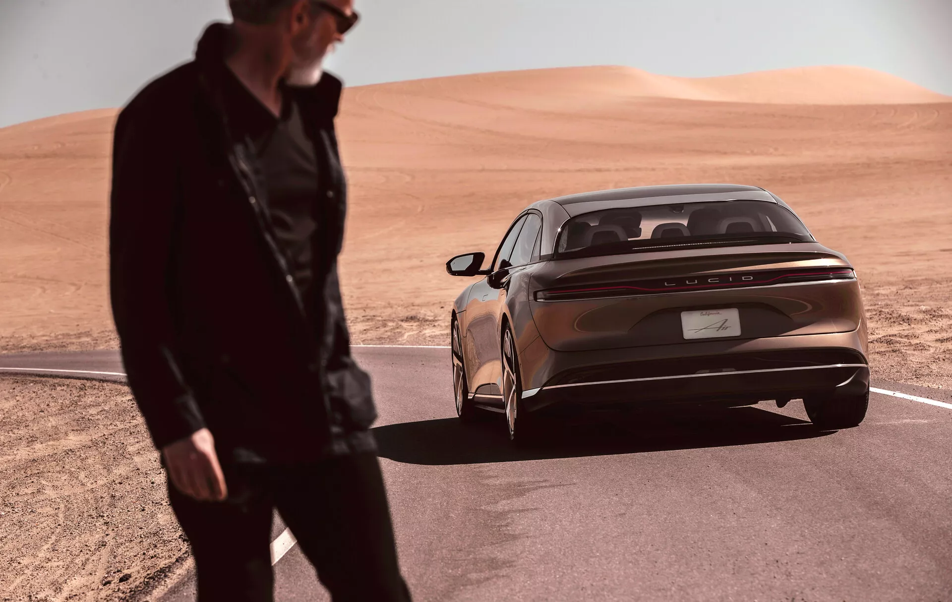 Una berlina Lucid Air 2022 (1ª Generazione) elegante e moderna parcheggiata in un paesaggio desertico, ripresa da un angolo basso con enfasi sui tre quarti posteriori. Il colore bronzo metallizzato della carrozzeria e le luci posteriori orizzontali prominenti sottolineano il design contemporaneo dell'auto. Una striscia cromata riflettente corre lungo il retro sotto le luci. Il badge del produttore è centrato sul cofano. Il paraurti posteriore integrato è dotato di sensori di parcheggio, aggiungendo al look lussuoso. Una figura sfocata è in primo piano, suggerendo movimento o attività vicino al veicolo.