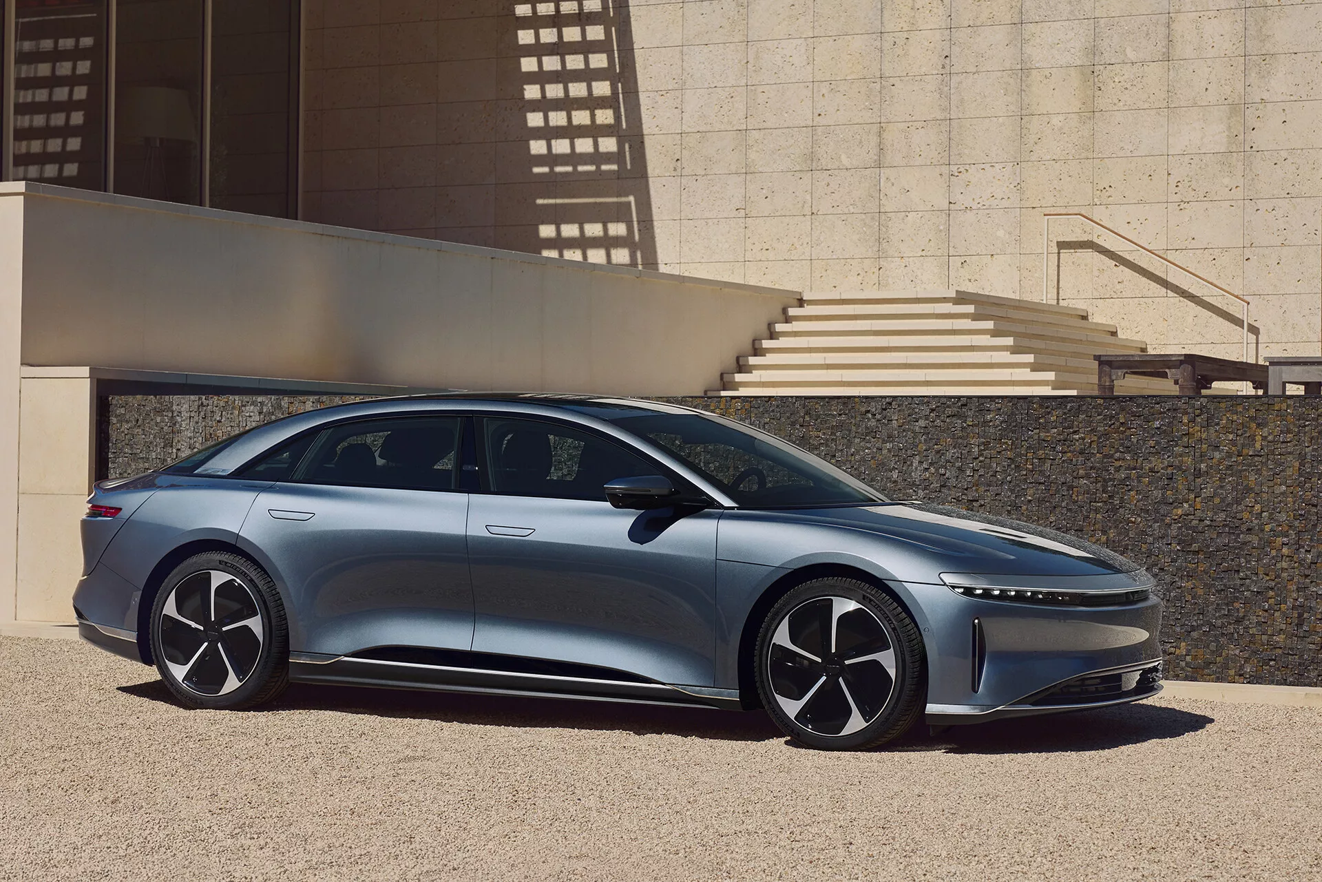 Un profilo laterale della Lucid Air 2022, una berlina moderna ed elegante, catturata all'aperto in un ambiente urbano con elementi architettonici che proiettano ombre. Il lato sinistro dell'auto mostra il suo design aerodinamico con una linea del tetto bassa, maniglie delle porte a filo, grandi ruote multirazze e specchietti laterali sottili. L'immagine mette in risalto la lunghezza dell'auto, dal paraurti anteriore a quello posteriore, con un montante A inclinato in avanti che conferisce un aspetto spazioso agli interni e una linea caratterizzante dinamica sulla carrozzeria.