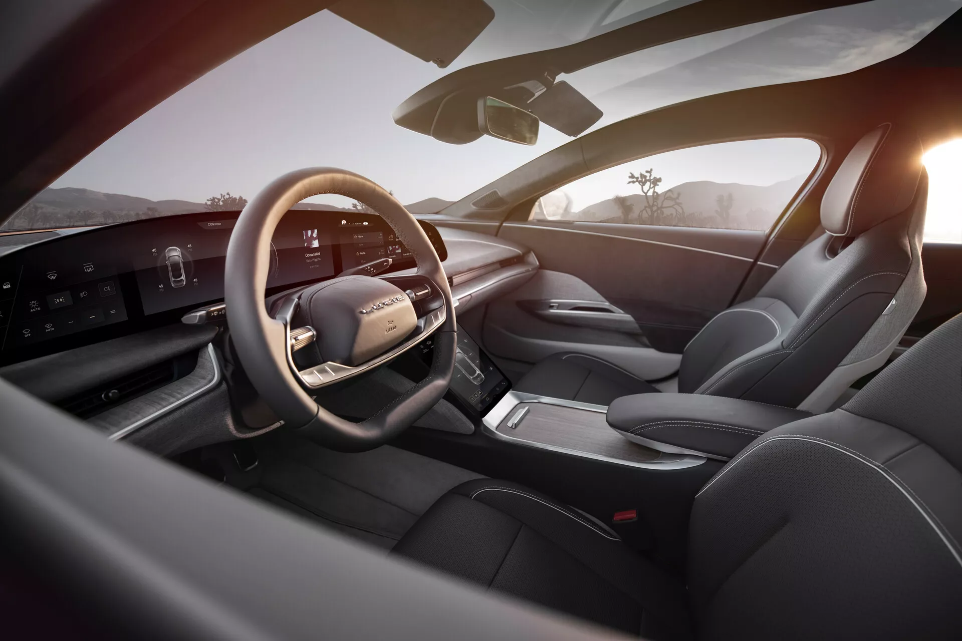 Vista interna della Lucid Air 2022 dalla prospettiva del sedile posteriore destro, mostrando un'area del conducente lussuosa e moderna. I punti salienti includono un volante multifunzionale rivestito in pelle con comandi integrati, un elegante display digitale orizzontale del cruscotto, una console centrale minimale con selettore di marcia elettronico, sedili sportivi rivestiti in pelle, sofisticati comandi del pannello porta e una buona visibilità generale con specchietto laterale e parabrezza. L'interno ben illuminato mette in risalto la tecnologia all'avanguardia e il design premium, accentuato dalla calda luce esterna che suggerisce alba o tramonto.