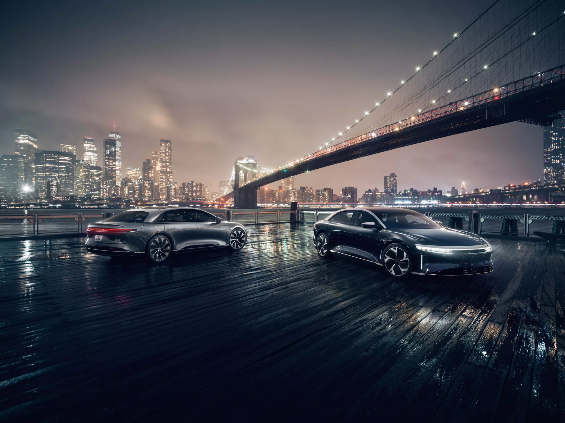 Due auto eleganti in un contesto urbano notturno con l'iconico Ponte di Brooklyn sullo sfondo. Le auto sono riprese da un'angolazione bassa, mettendo in risalto i loro dettagli di design. L'auto in primo piano, angolata verso la telecamera, mostra la griglia frontale, i fari a LED e i tergicristalli con sensore pioggia. L'auto in secondo piano, parallela alla telecamera, rivela il suo profilo laterale, le ruote in lega e la verniciatura di alta qualità. Entrambi i veicoli sono su una superficie bagnata che riflette l'illuminazione a LED, suggerendo tecnologie avanzate come l'ABS e il controllo di trazione.