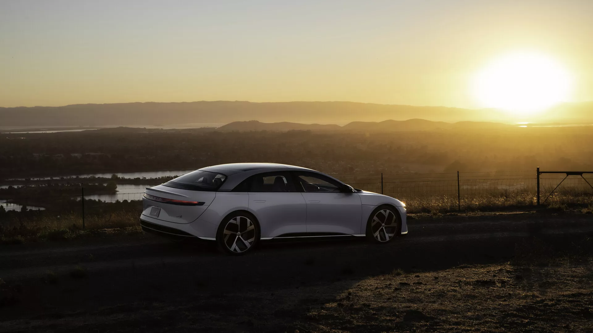 Lucid Air 2022 (1ª Generazione) fotografato da un'angolazione laterale posteriore durante l'alba o il tramonto con un sole caldo sullo sfondo, posizionato sul lato sinistro dell'inquadratura. La berlina a quattro porte, elegante e moderna, presenta linee fluide, grandi ruote multirazza e maniglie delle portiere retrattili. Il paesaggio comprende corpi d'acqua e un punto di osservazione elevato. Le luci posteriori sono strette e allungate orizzontalmente, con la luce del sole che crea un effetto di bagliore caldo e flare nell'immagine.