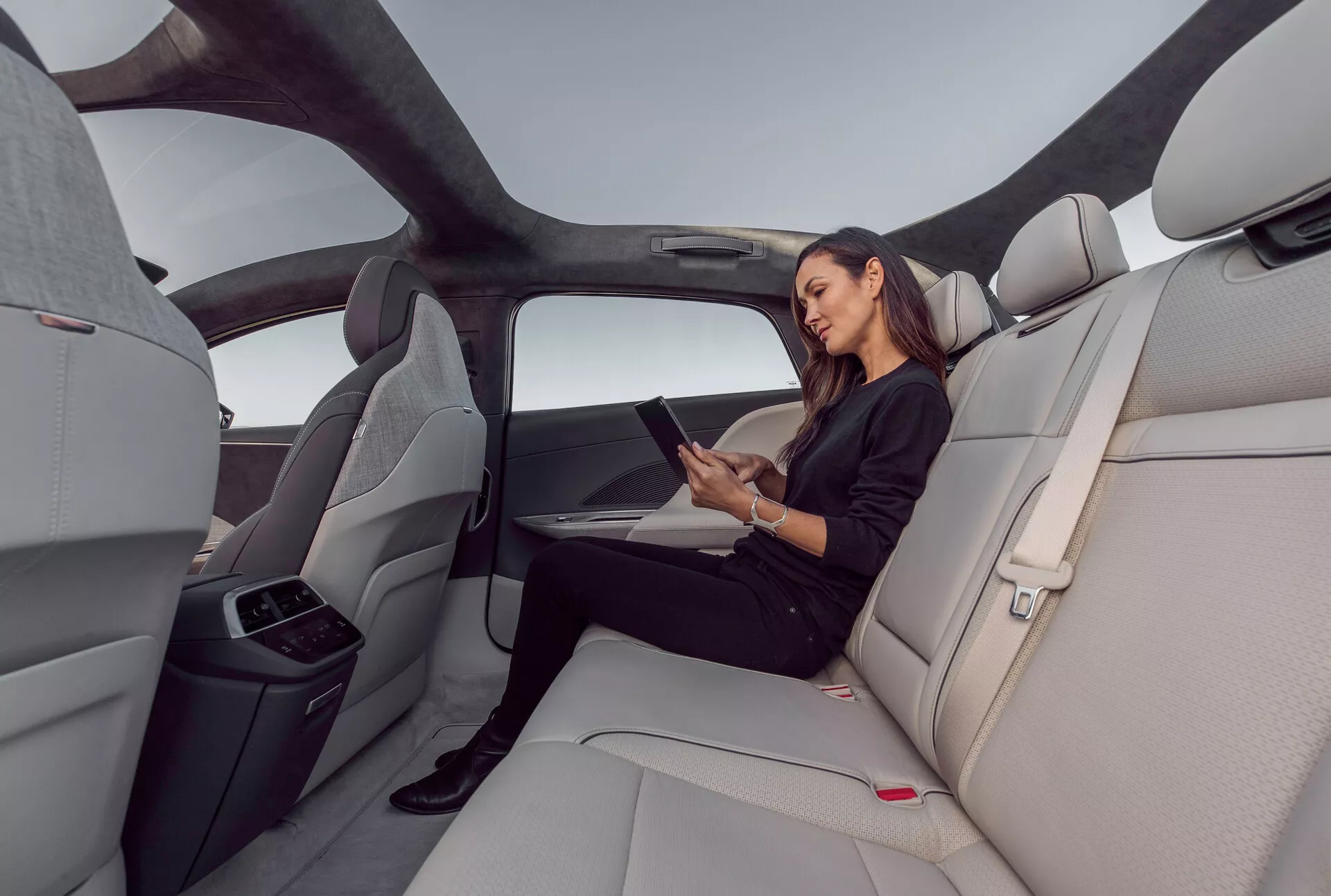Vista interna dell'area di seduta posteriore della Lucid Air 2022, evidenziando la spaziosa cabina di lusso con sedili sagomati in pelle grigio chiaro. L'immagine cattura una ripresa grandangolare dal lato destro, mostrando il tetto panoramico che si estende sopra l'area posteriore, design ergonomici dei sedili e un bracciolo centrale piegato con portabicchieri. Una donna è seduta comodamente interagendo con un tablet, indicando possibili funzionalità di connettività. Interno elegante e minimalista con bottoni di rilascio della cintura di sicurezza rossi e linee orizzontali sul cruscotto e sui pannelli delle porte, migliorando la sensazione di larghezza e apertura.