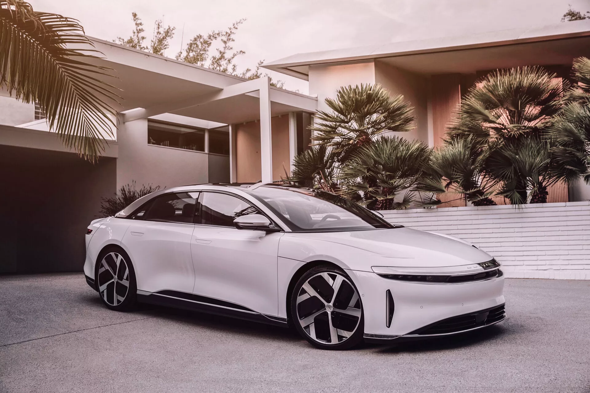 L'immagine raffigura una Lucid Air 2022 di prima generazione, una berlina bianca moderna ed elegante con assetto basso e ampio e linea del tetto simile a una coupé. Catturato da un'angolazione bassa, questo scatto accentua il suo design aerodinamico, i grandi cerchi distintivi, i fari a LED e gli specchietti laterali aerodinamici, suggerendo tecnologie avanzate di assistenza alla guida. La griglia anteriore minimalista suggerisce tecnologie di propulsione elettrica o ibrida. Il design futuristico dei cerchi presenta coperture aerodinamiche e l'aspetto del veicolo è valorizzato da finestrini oscurati, porte senza telaio e maniglie a filo eleganti. Una vista parziale del tetto panoramico suggerisce un'abitacolo lussuoso e spazioso. Lo sfondo mostra un'area residenziale esclusiva con palme, indicando che l'auto è destinata ai consumatori benestanti.