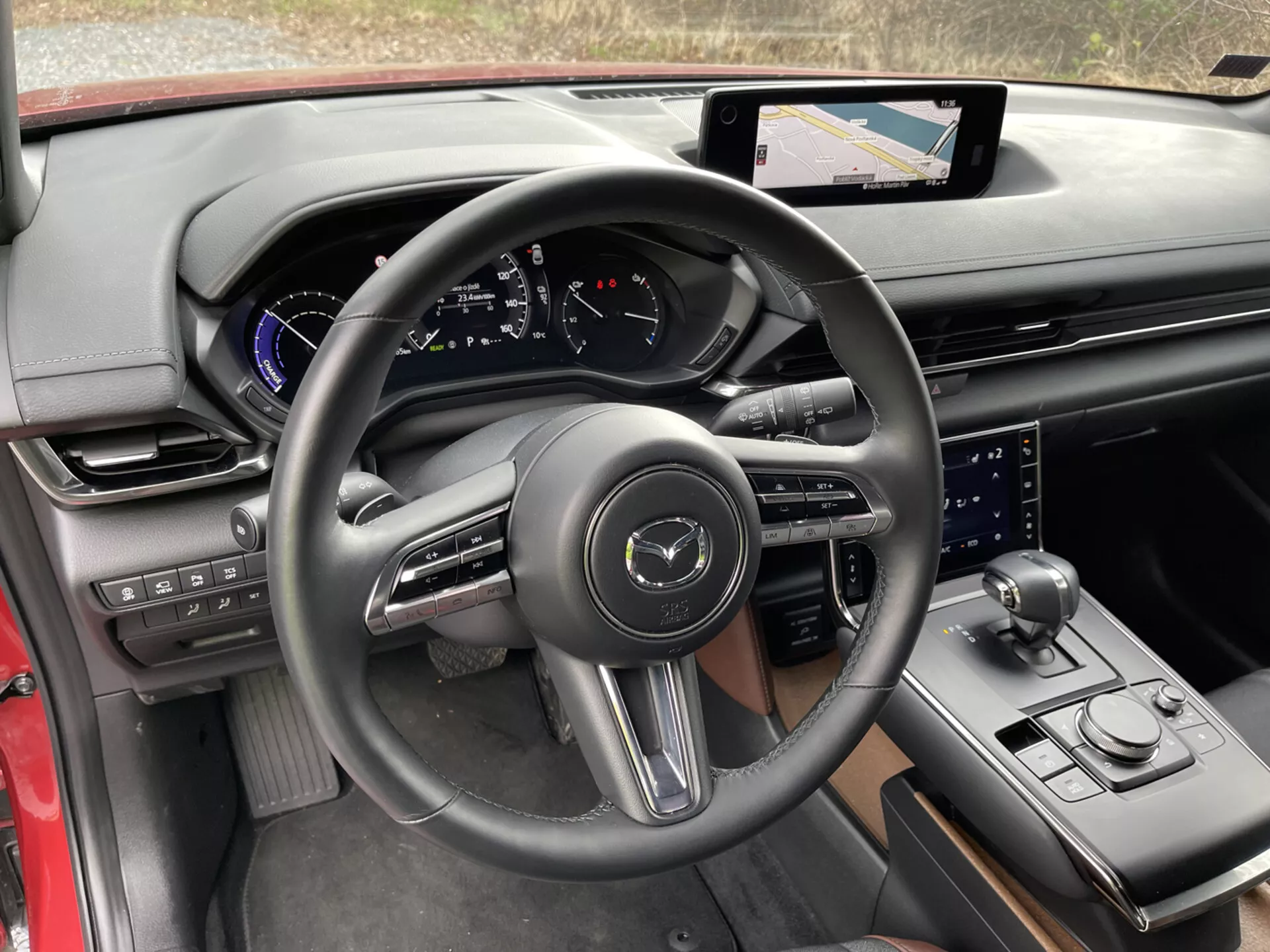 Vista interna della Mazda MX-30 2020 dalla prospettiva del conducente, con focus sui dettagli quali il volante a tre razze avvolto in pelle con pulsanti di controllo e paddle shift, cluster strumenti digitale sul cruscotto con accenti blu, sistema di infotainment con touch screen e navigazione sulla console centrale, leva del cambio sportiva, selettore della modalità di guida, ulteriore manopola di controllo per il sistema di infotainment, mix lussuoso di materiali con finiture nere e rosse, portabicchieri, e pannelli di controllo sulla porta per specchietti laterali e finestre, evidenziando la tecnologia moderna e il design ergonomico.